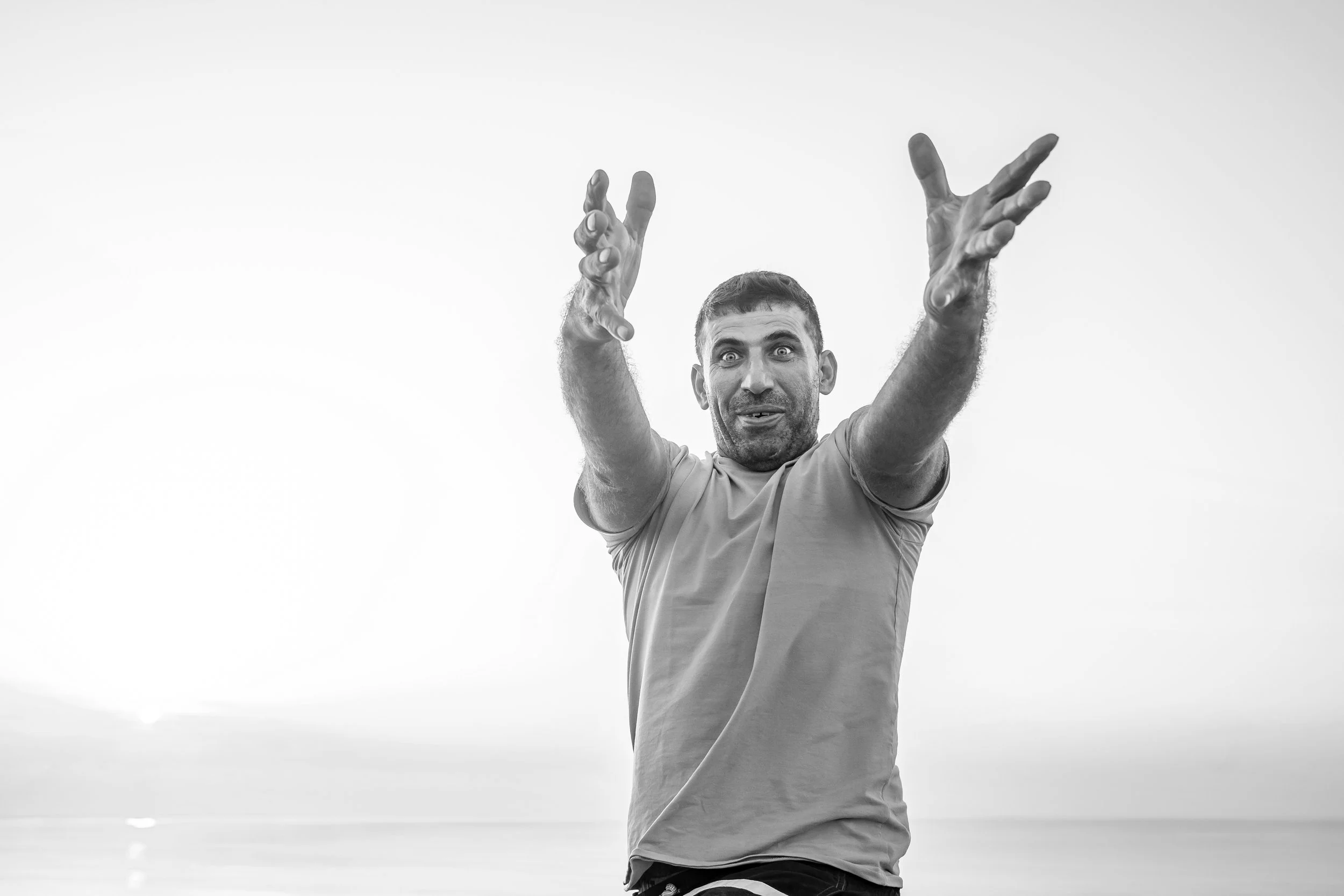 Mann mit kurzen dunklen Haaren und Bart, der vor einem weißen Himmel steht, mit ausgebreiteten Armen und einem überraschenden oder enthusiastischen Ausdruck.