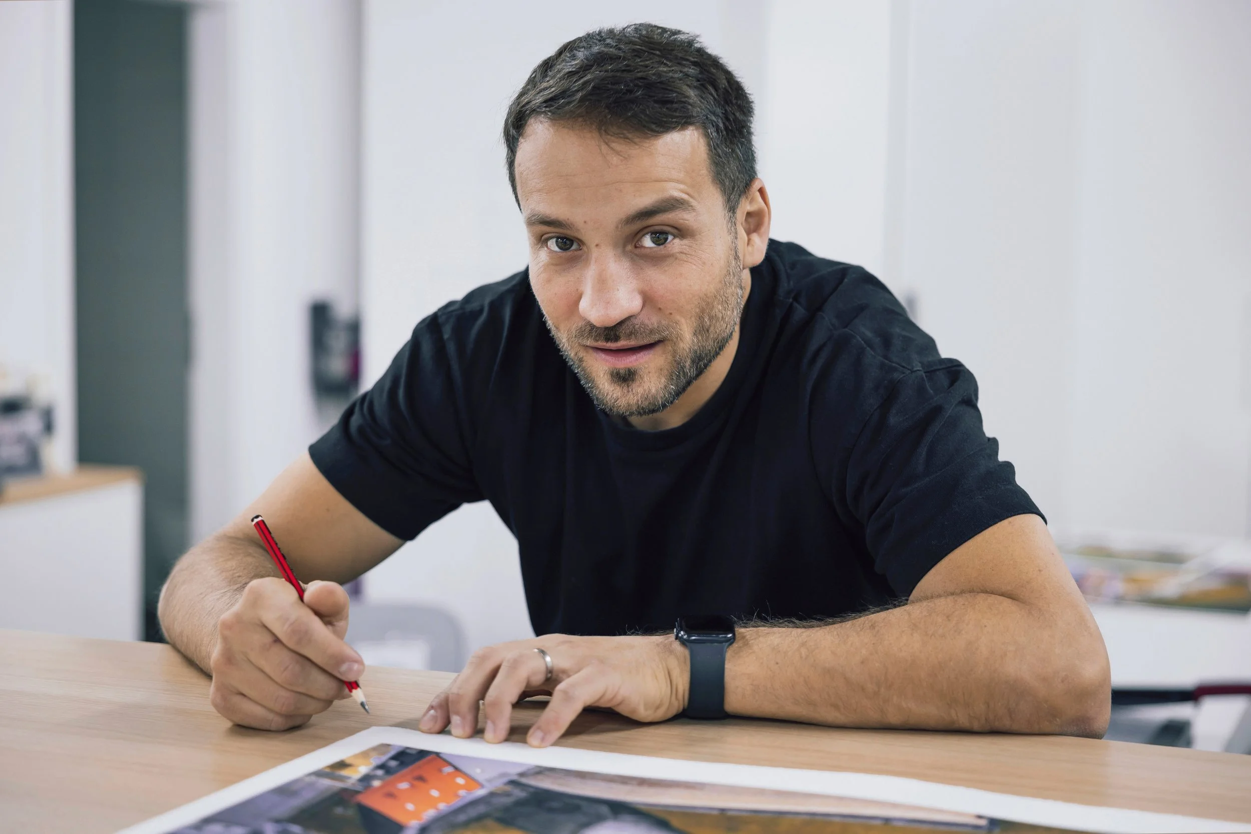 Mann mit dunklem T-Shirt, braune Haare und Bart sitzt an einem Tisch, hält einen roten Stift und betrachtet eine fotografische Skizze.