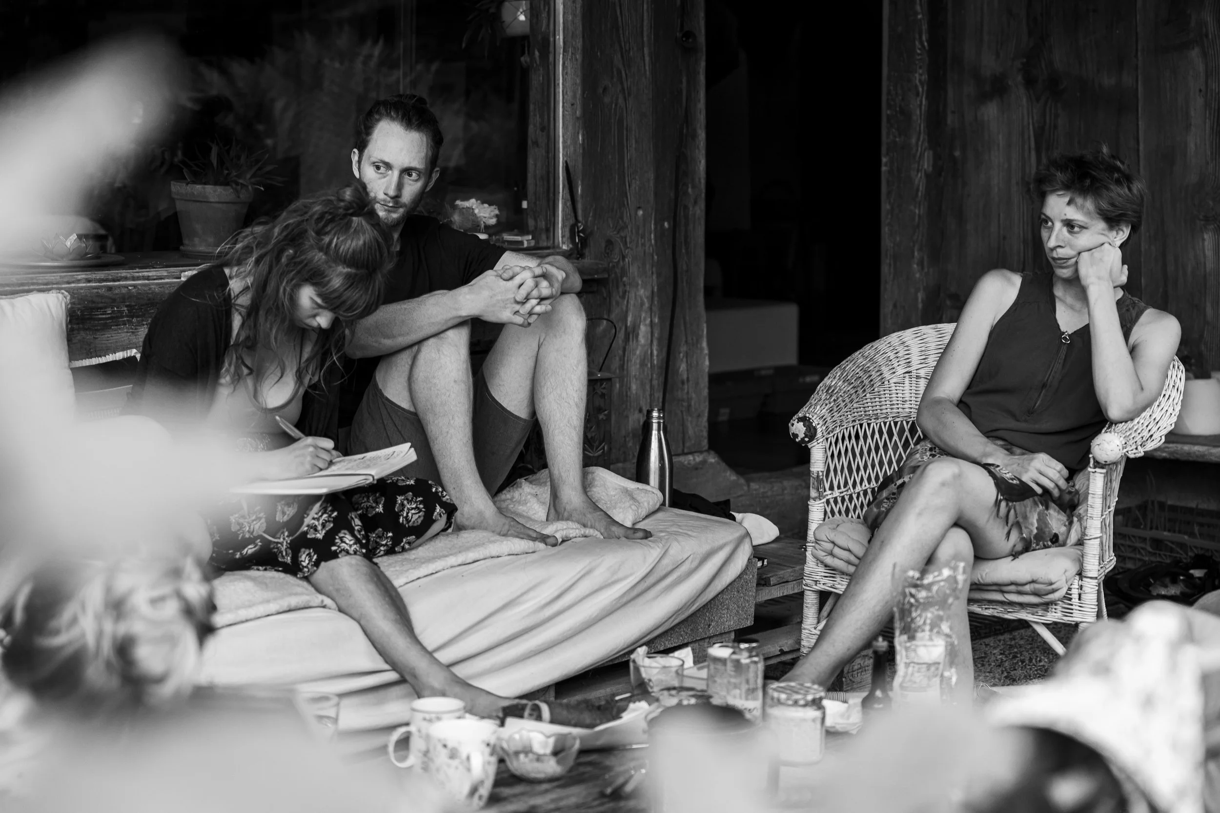 Vier Menschen sitzen in einem gemütlichen Raum. Zwei Menschen sitzen auf einer Couch, eine Person schreibt in ein Notizbuch, während die andere Person daneben sitzt. Ein weiterer Mensch sitzt in einem Korbstuhl auf der rechten Seite, schaut nachdenkl