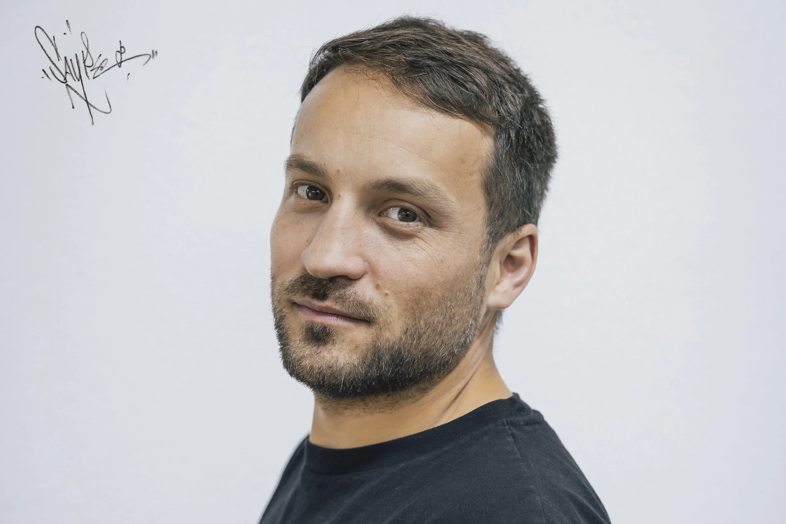 Nahaufnahme eines Mannes mit dunklem kurzen Haar, Bart und schwarzem T-Shirt, vor weißer Wand im Hintergrund.