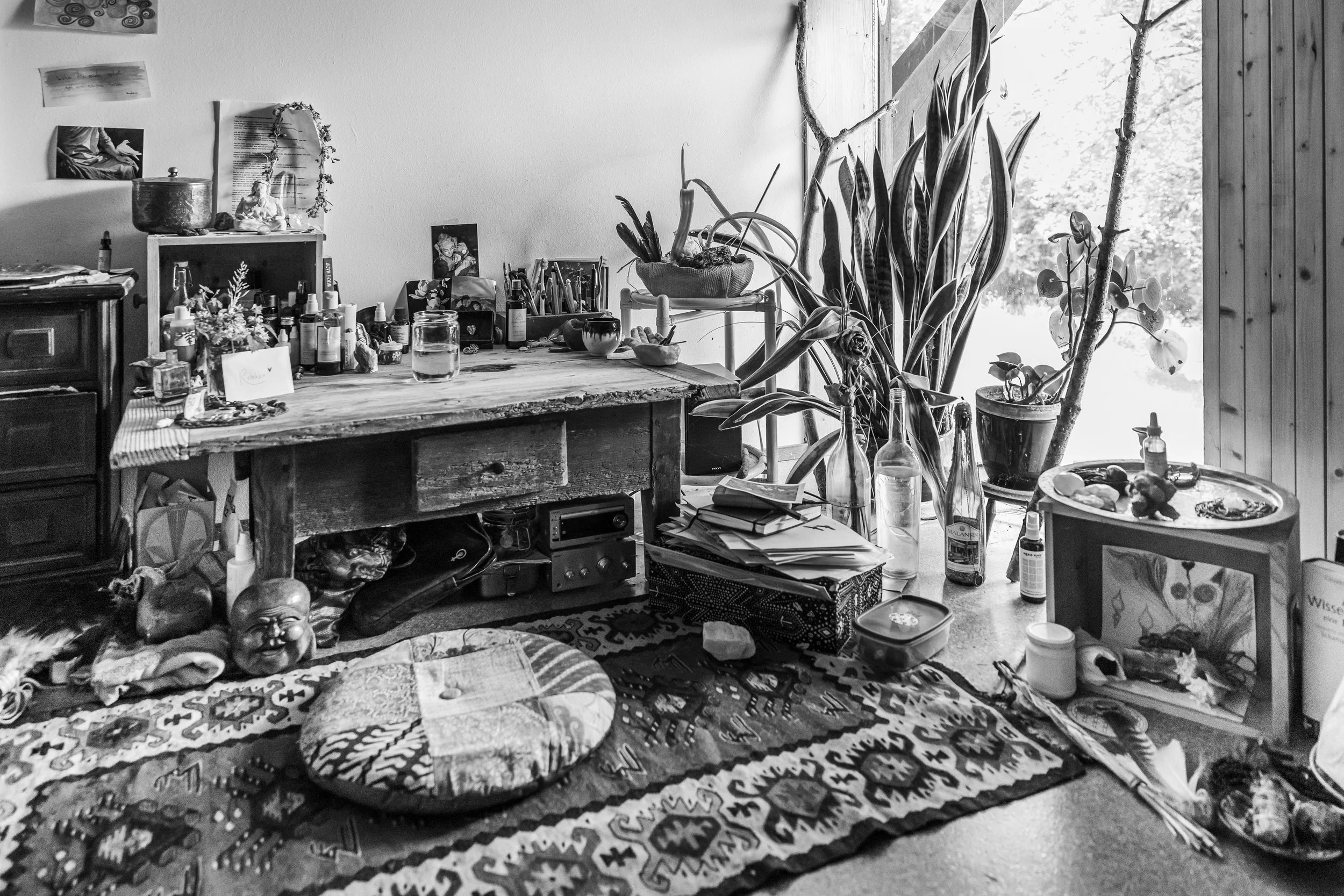 Chaotisch eingerichteter Raum mit einem alten Holzschreibtisch, verschiedenen Pflanzen, Flaschen und persönlichen Gegenständen, auf einem stilvollen Teppich.