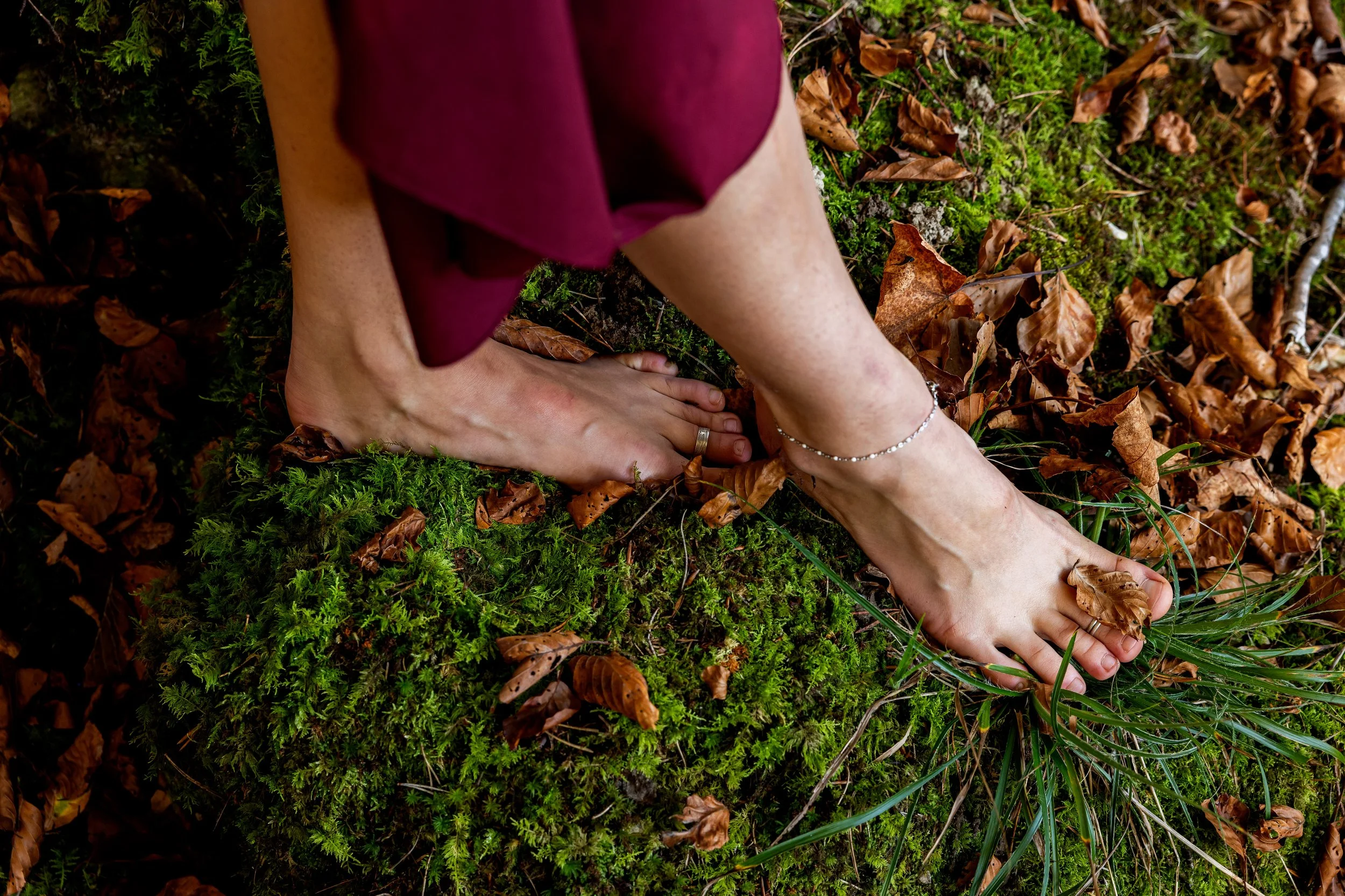 Eine Person steht barfuß auf Moos und Blätter im Wald, trägt eine silberne Kette und einen Ring am Finger.