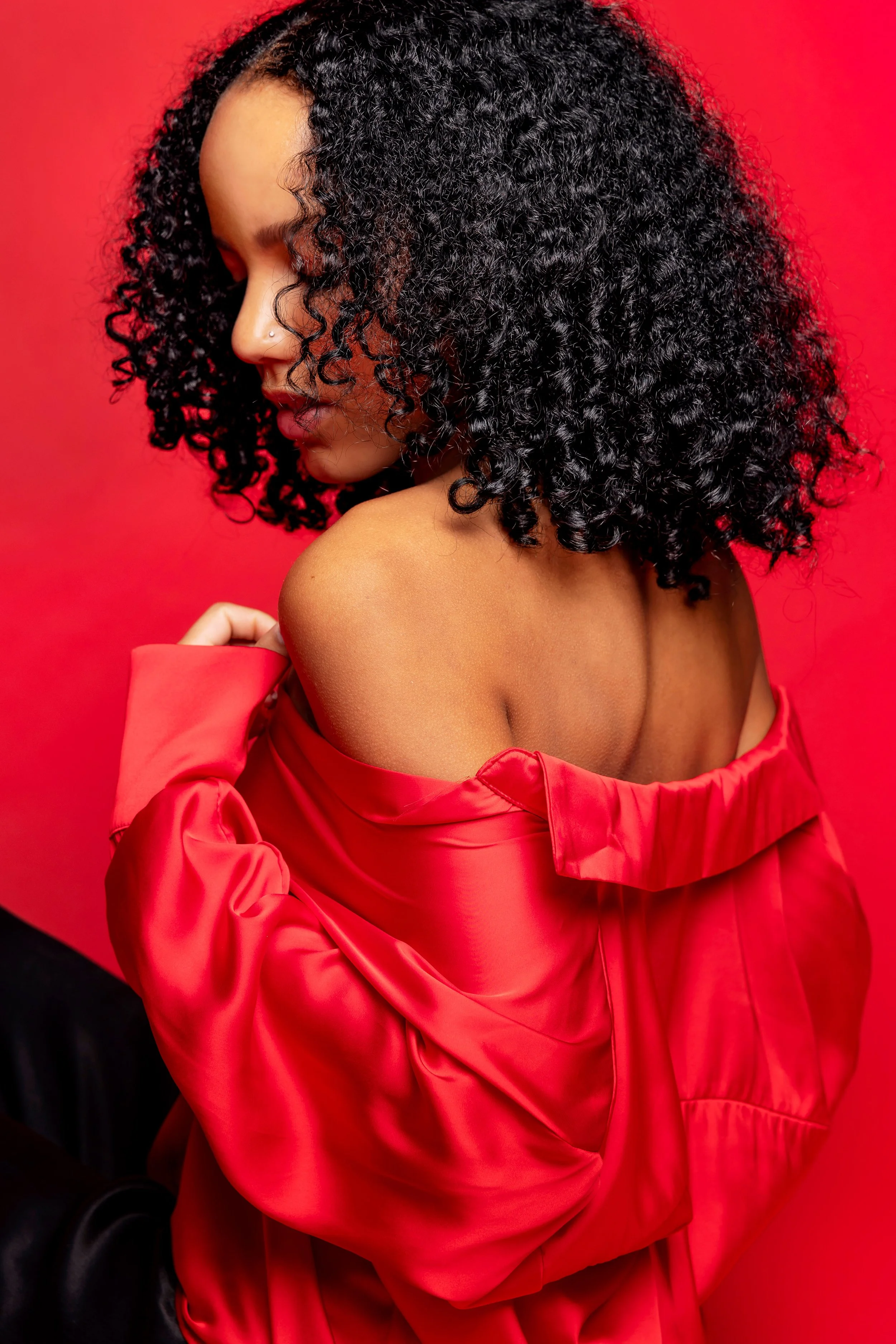 Eine Frau mit lockigem schwarzen Haar, die ein rotes Satin-Kleid trägt und seitlich fotografiert ist, vor einem roten Hintergrund.