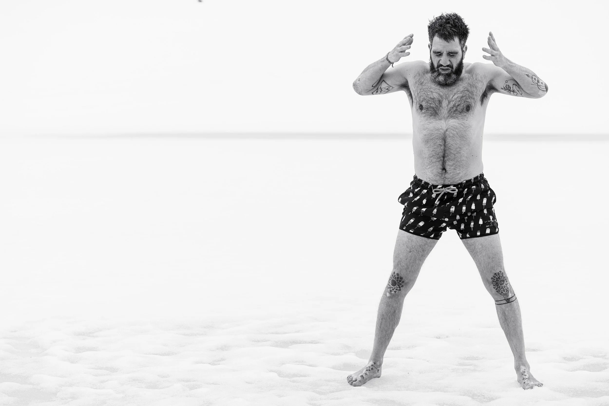 Ein nackter Mann mit Tattoos steht auf einer weißen, leeren Fläche, vermutlich Salz oder Sand, mit verschränkten Armen und einem nachdenklichen Gesichtsausdruck.