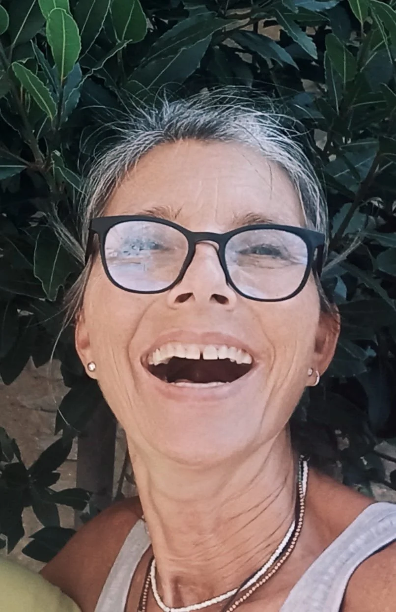 A woman with gray hair and glasses smiling widely in front of green leafy bushes.