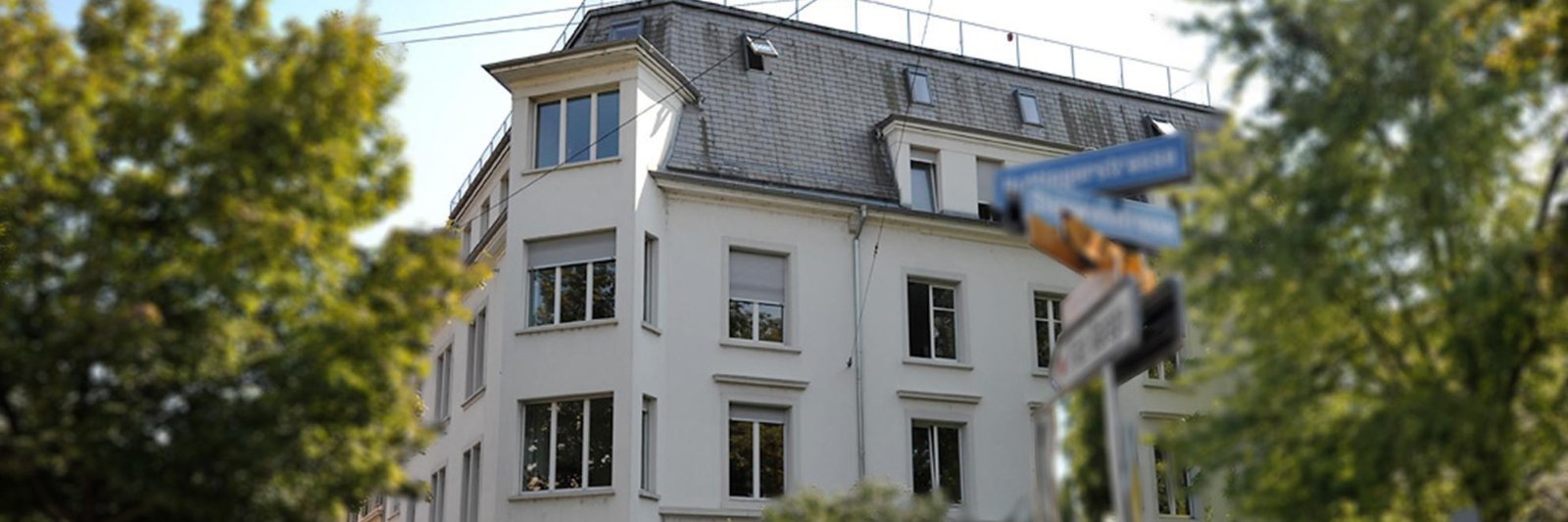 Ein mehrstöckiges weißes Wohnhaus mit einer Schieferdach und mehreren Fenstern, umgeben von grünen Bäumen, mit einem Straßenschild im Vordergrund.