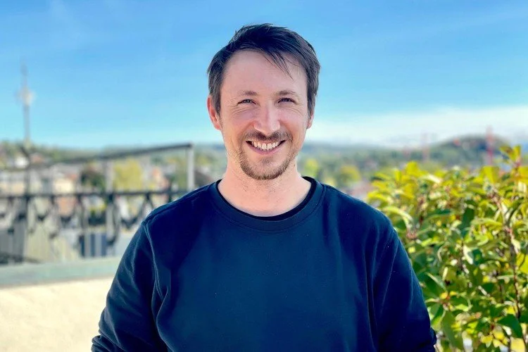 Ein Mann mit dunklem Haar und Bart steht auf einem Balkon, im Hintergrund eine grüne Landschaft und ein blauer Himmel.
