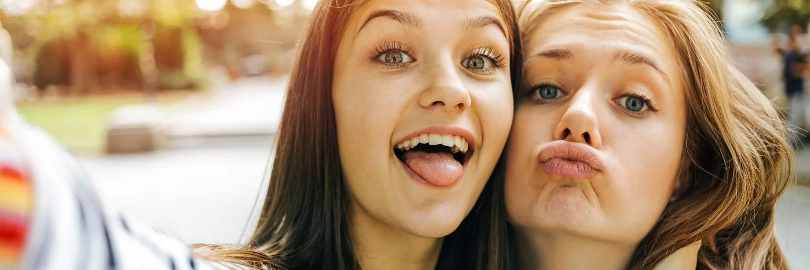 Zwei junge Frauen machen ein Selfie im Freien, eine lächelnd mit Zunge heraus, die andere pfeifend.