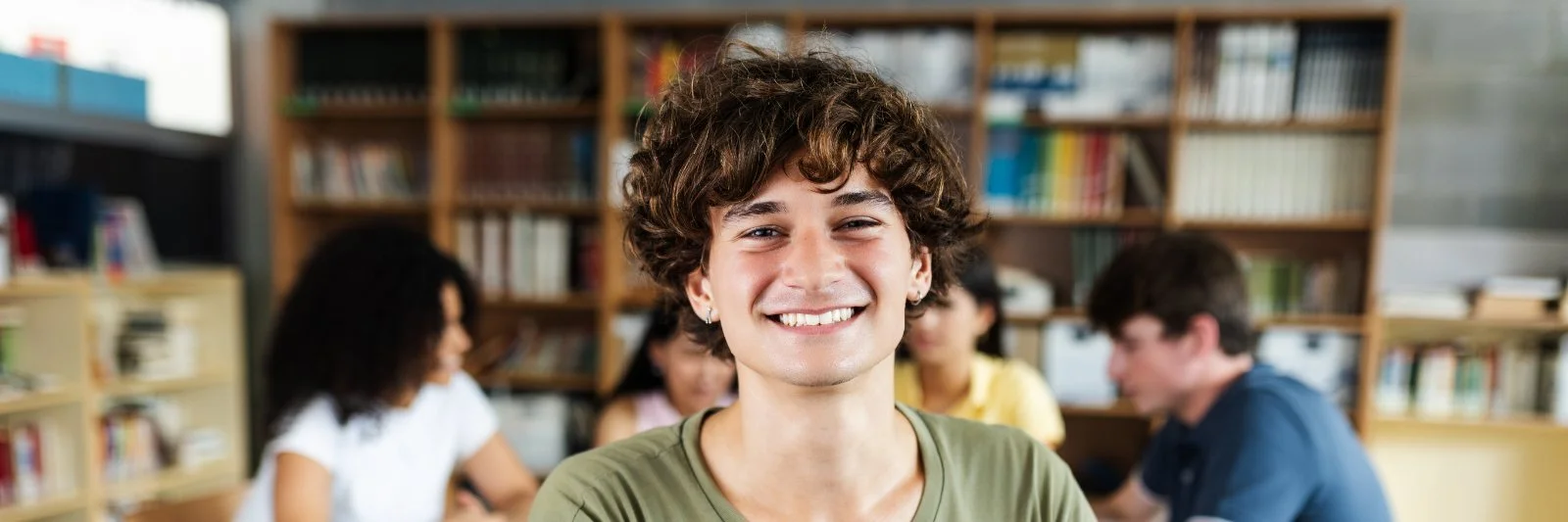 Eine junge Person mit kurzen, lockigen Haaren lächelt in einem Raum mit Bücherregalen, im Hintergrund sitzen andere Jugendliche bei einem Gruppentreffen.