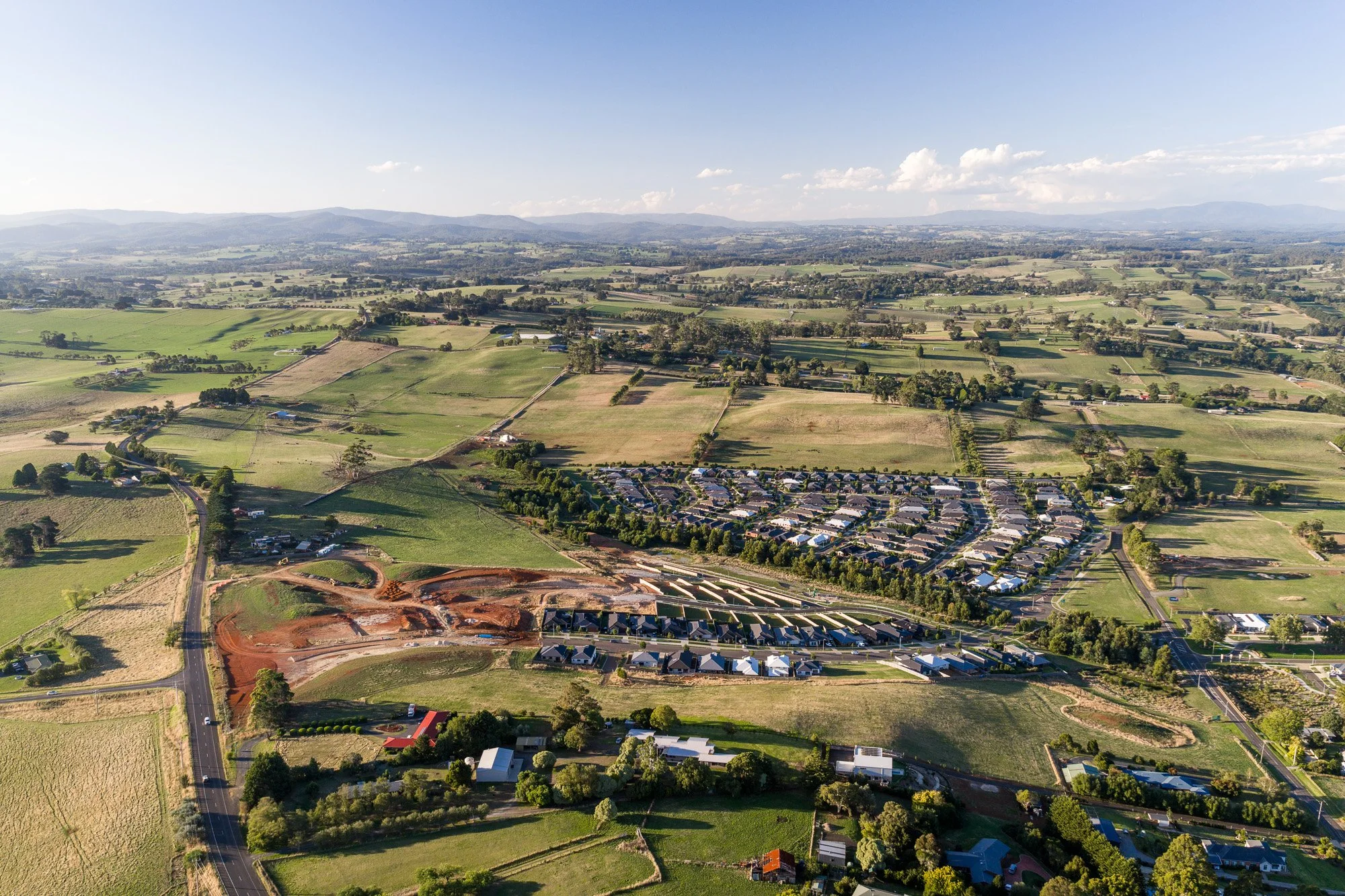 Aerial view of a construction site with reddish dirt, some houses, and a neighborhood surrounded by green fields and farmland.
