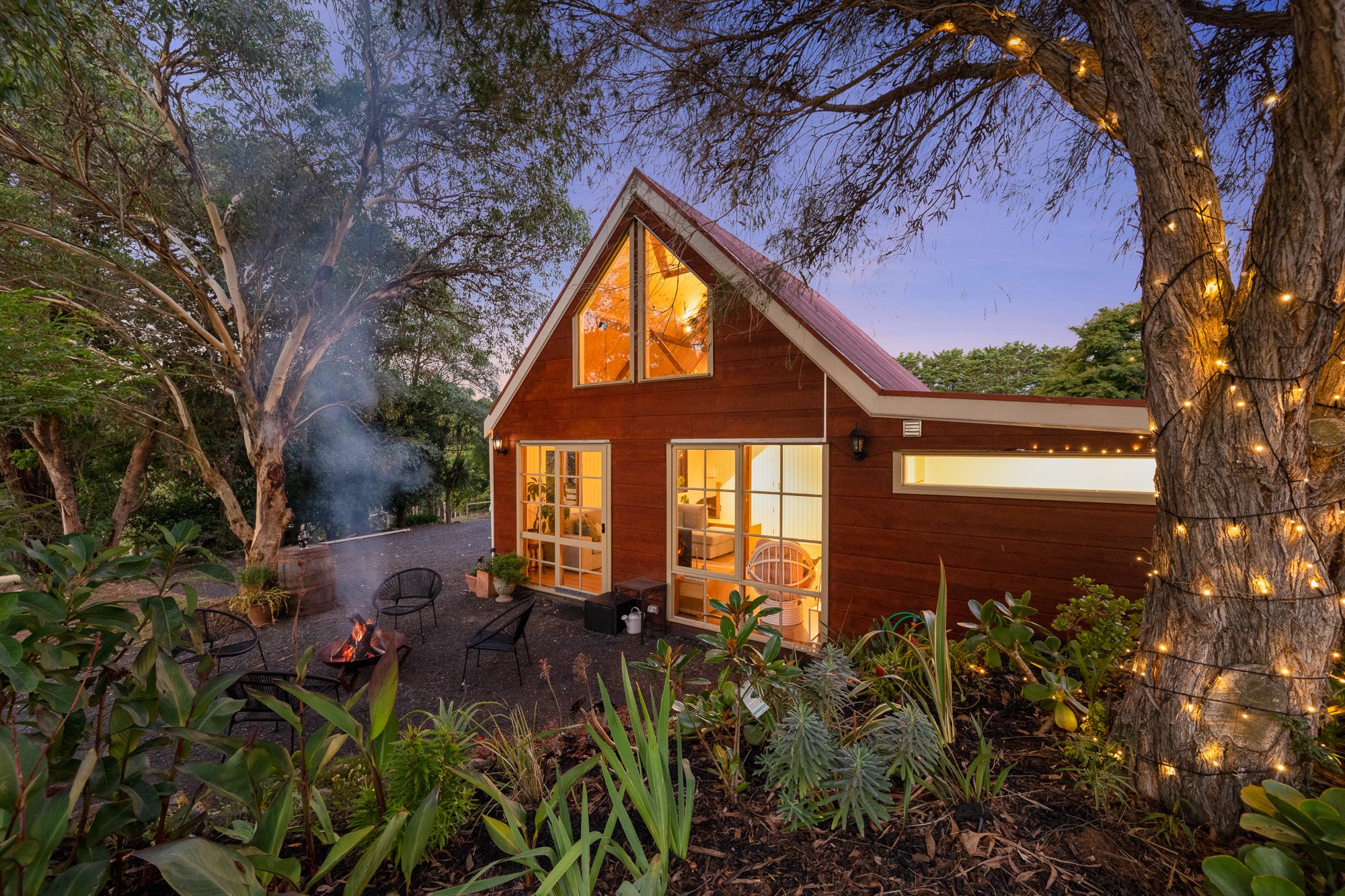 A cozy wooden house with large glass windows and a red metal roof, surrounded by lush greenery, at dusk. The house has interior lights on, and outside there is a small patio with chairs and a firepit, with string lights wrapped around a large tree.