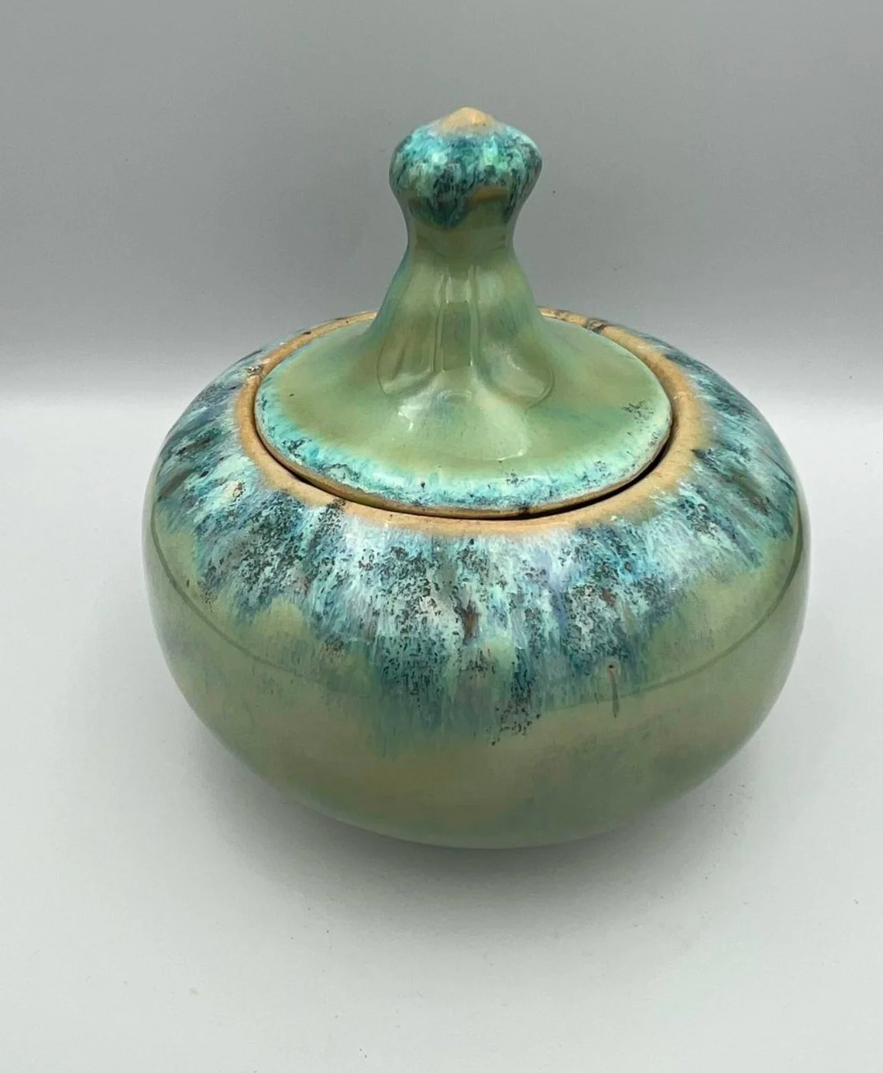 Een ronde keramieken urn met een deksel, glanzend groen-blauw glazuur met creatieve textuur en details.