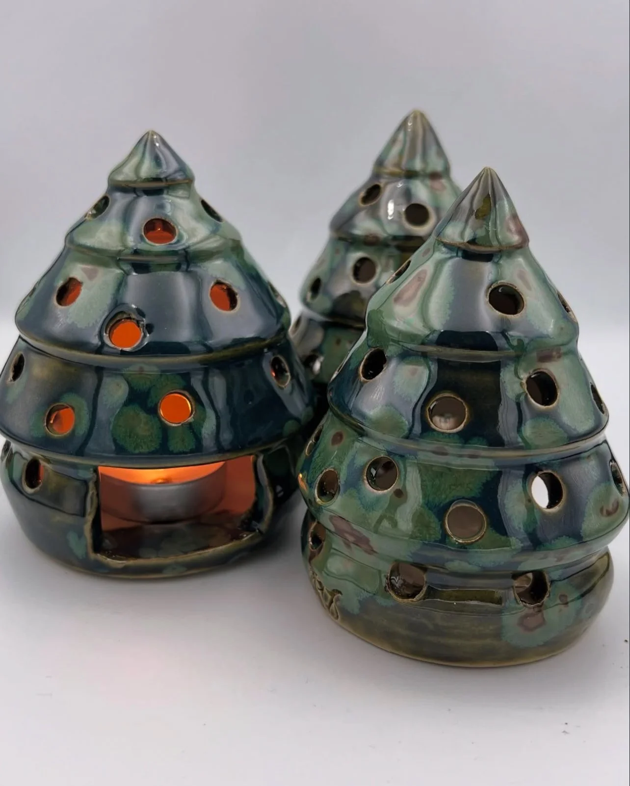 Drie keramische theelichthouders in de vorm van kleine kerstbomen met perforaties, waarvan één met een brandend lichtje erbinnen.