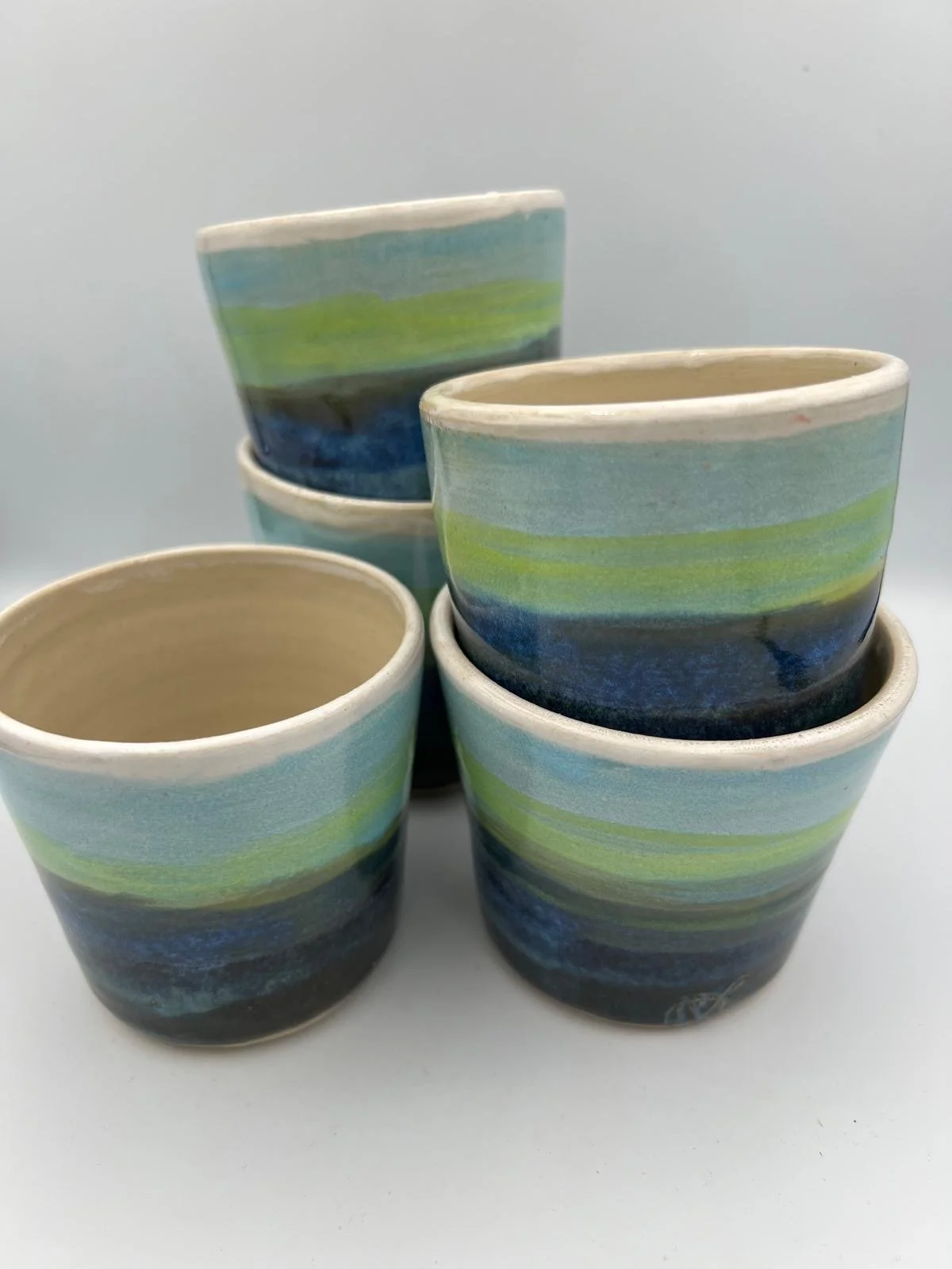 Vijf handgeschilderde keramische potten met blauwe, groene en zwarte strepen, gestapeld en gerangschikt op een lichte ondergrond.