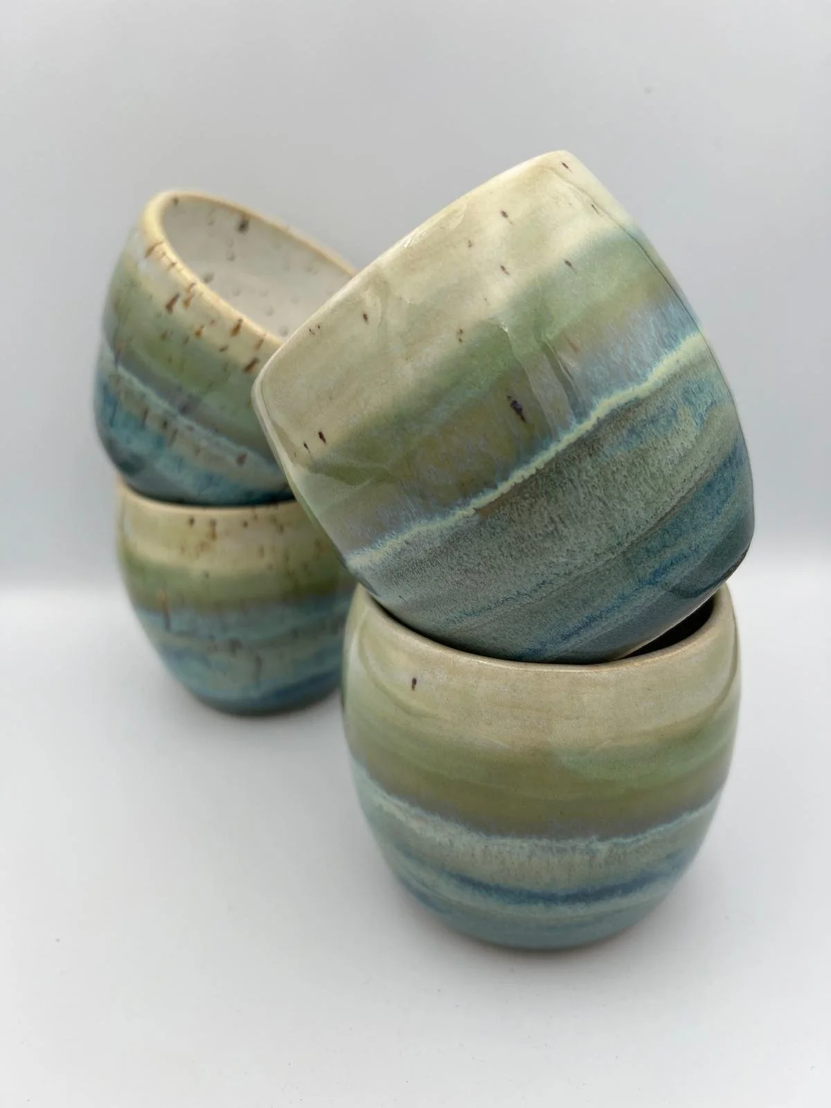 Vier kleine, gedraaide keramiekcupjes in pastelkleuren met blauwe, groene en beige tinten, gestapeld op een witte achtergrond.