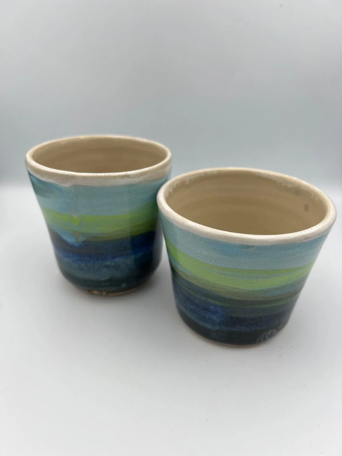 Twee handgemaakte keramische bekers met blauwe en groene strepen, wit binnenvlak op een witte achtergrond.