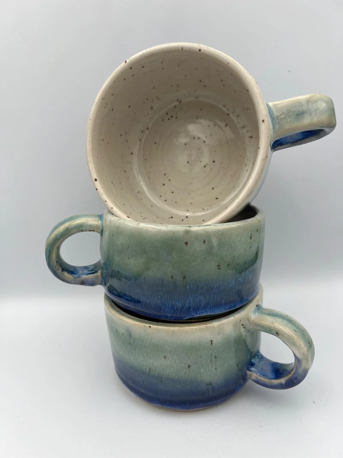 Twee gestapelde keramieken bekers met een blauwe en groene glazuurafwerking, één beker geplaatst op de ander, beiden met handvatten.