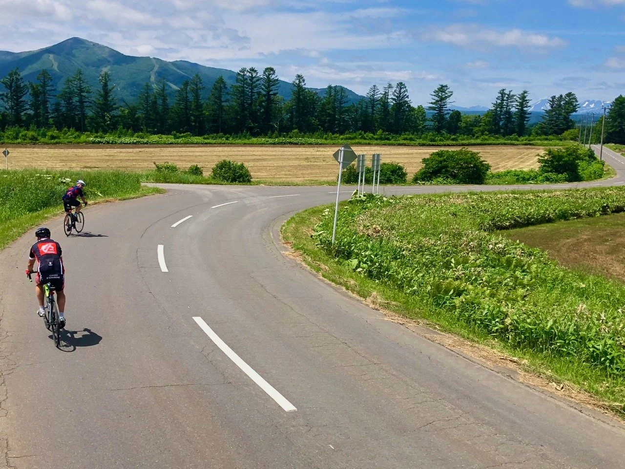 Two cyclists riding on a winding country road surrounded by lush green fields, trees, and mountainous landscape in the background.