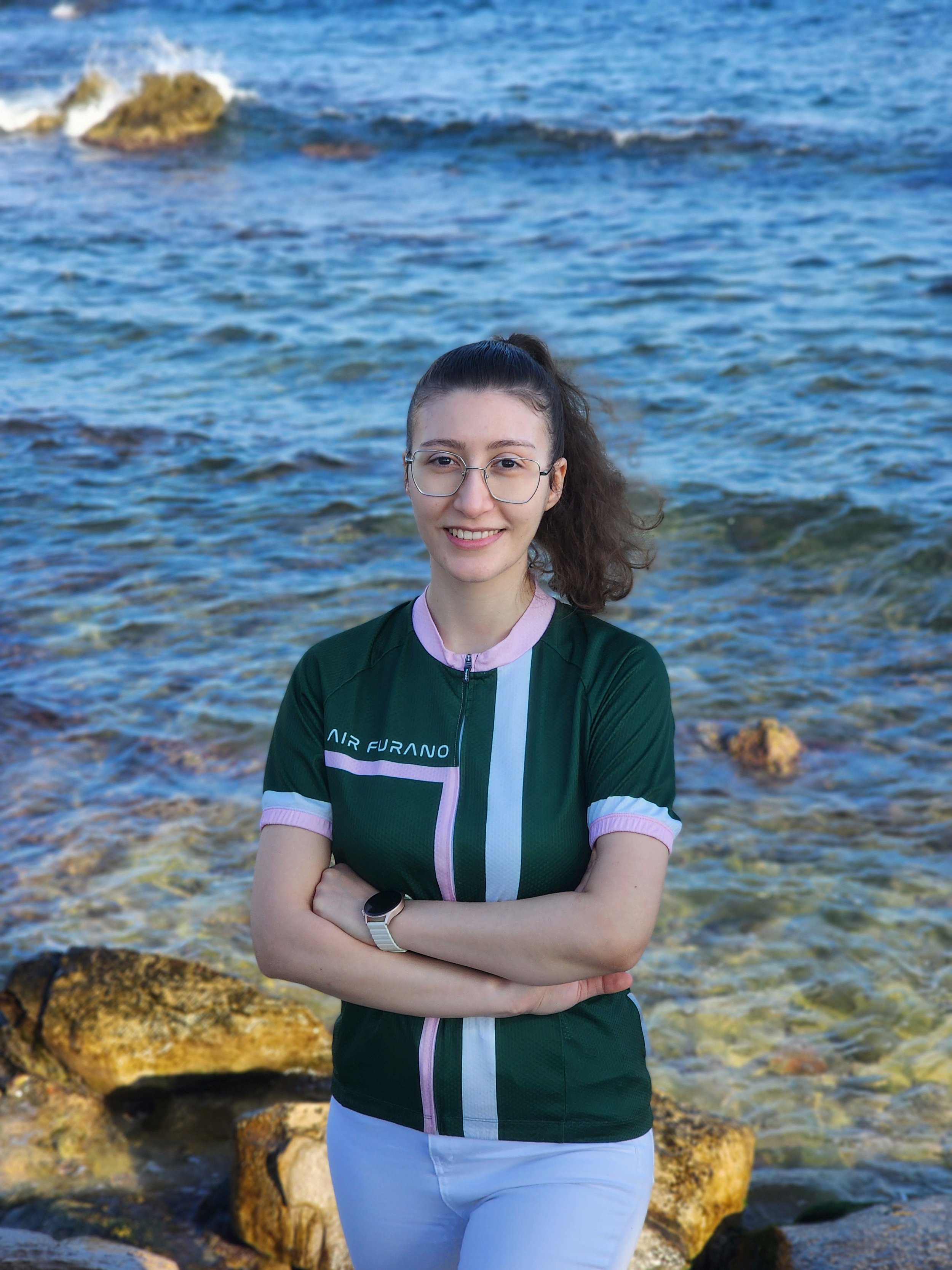 A young woman with brown hair, glasses, and a slight smile wearing a green and pink cycling jersey, by the beach.