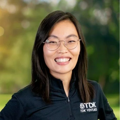 A woman with black hair and glasses smiling outdoors, wearing a black jacket with TDK Ventures logo.