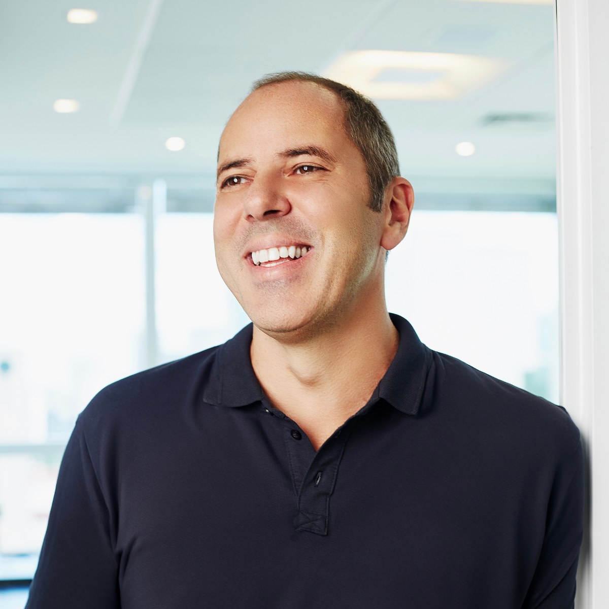 A smiling man with short hair, wearing a dark polo shirt, standing in a bright office with large windows.