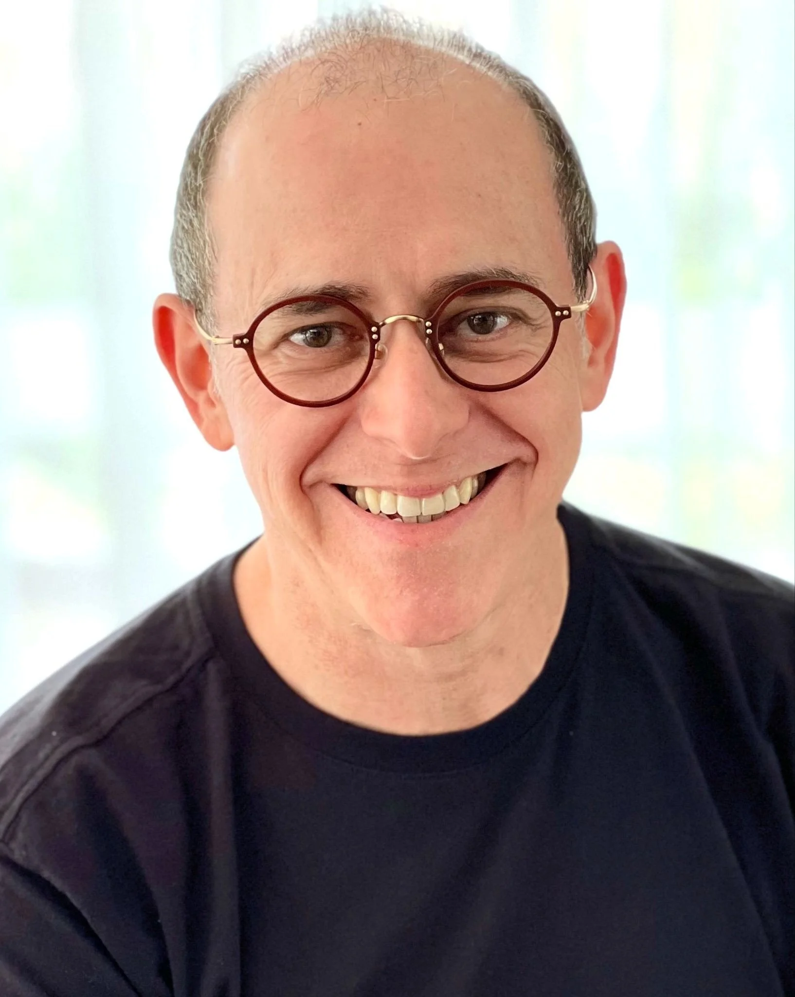 Close-up of a smiling person with glasses, short hair, and wearing a dark shirt.