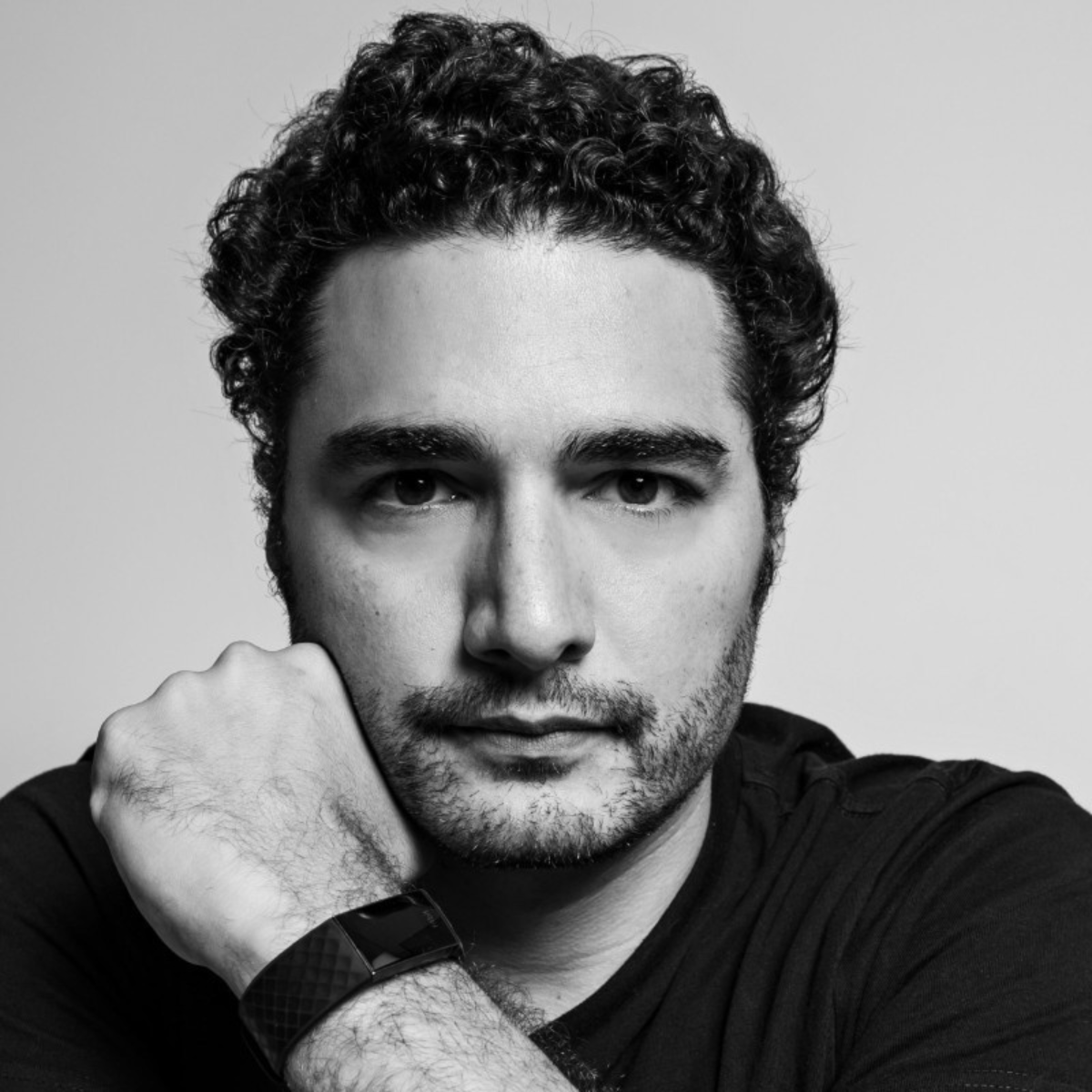 Black and white portrait of a young man with curly hair, resting his chin on his hand, wearing a watch, looking directly at the camera.