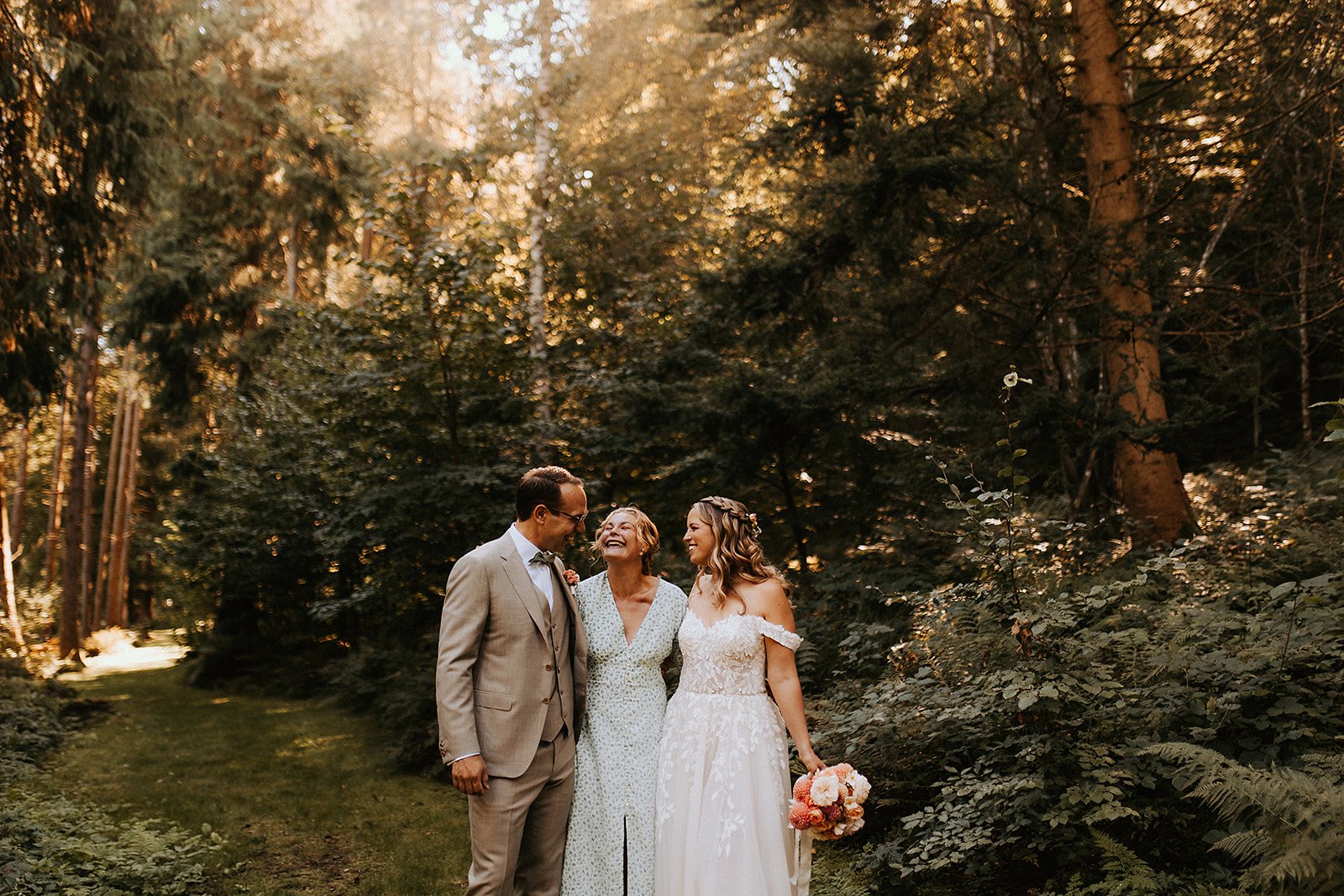A bride and groom standing outdoors, smiling and laughing with an older woman, among tall trees and lush greenery during a wedding celebration.