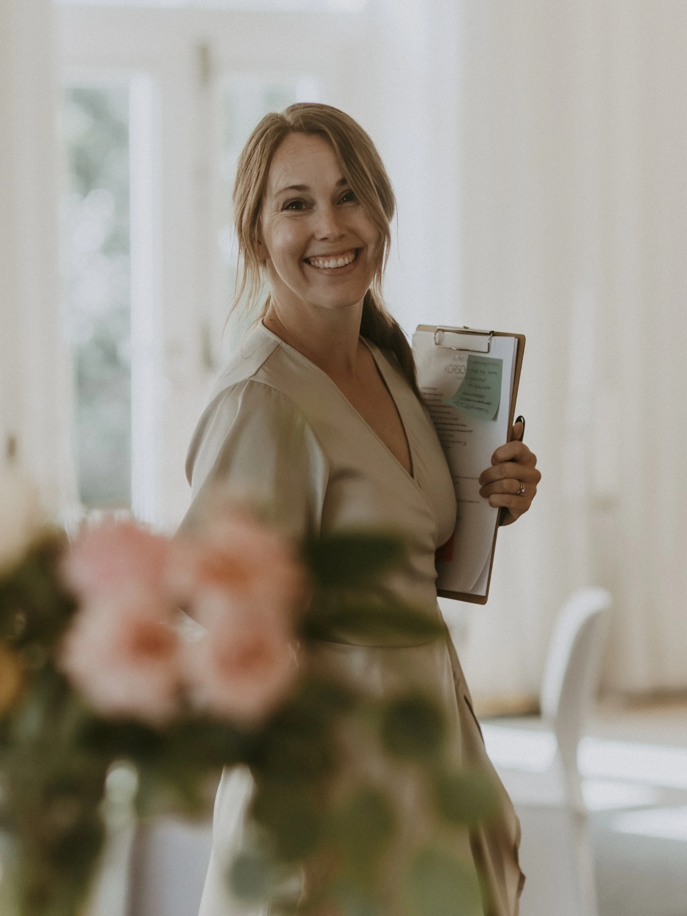 Smiling woman with light brown hair holding a clipboard in a bright white room.