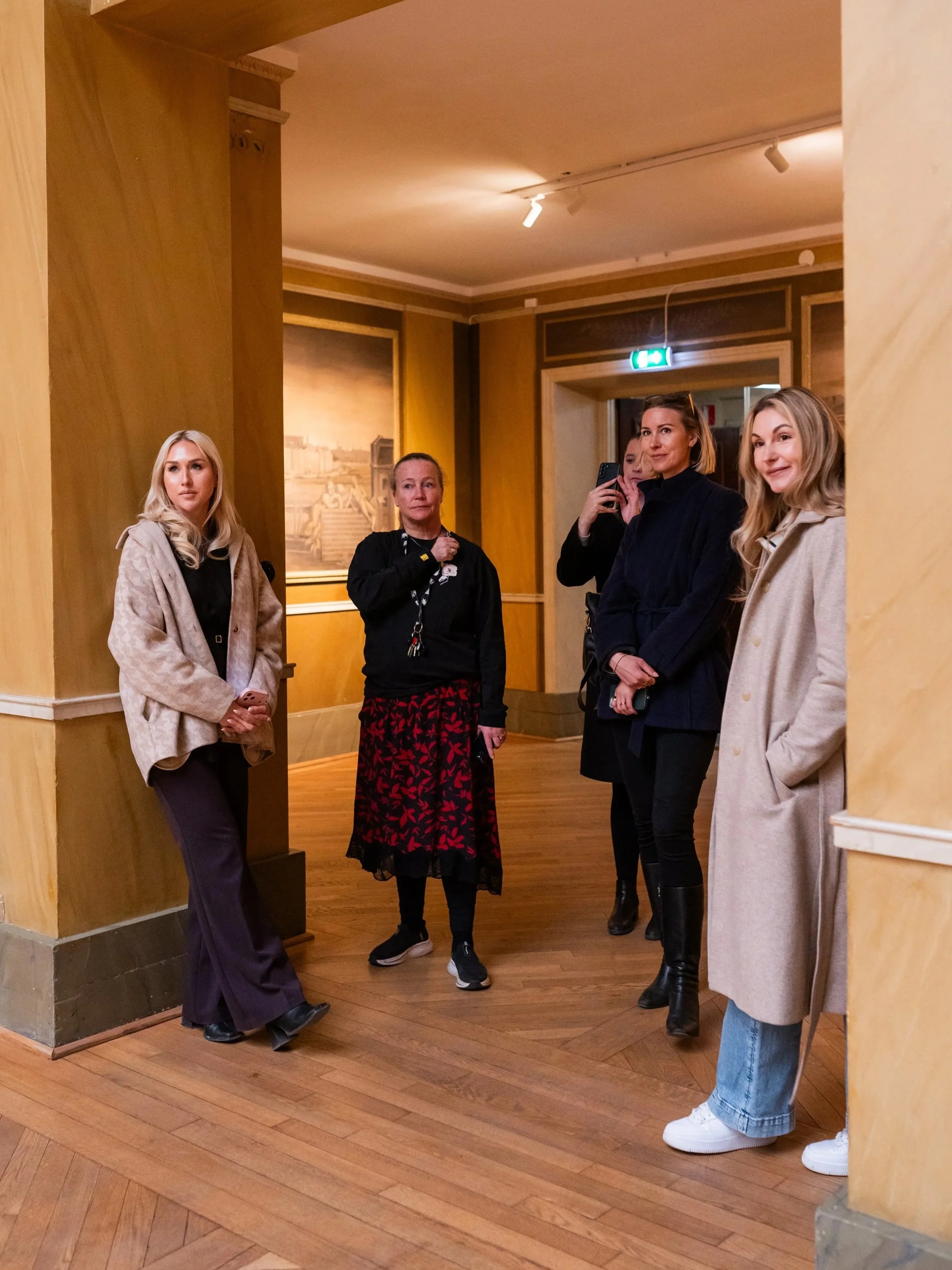 Five women standing and waiting in a room with wooden floors and tan walls. One woman on the far left is leaning against a wall, wearing a beige jacket and black pants. The second from the left is wearing a darker top and red patterned skirt, holding keys. The woman in the middle is taking a photo with a phone, dressed in black. The second from the right is wearing a dark coat and has blonde hair. The woman on the far right is wearing a beige coat and jeans, with part of her face visible.