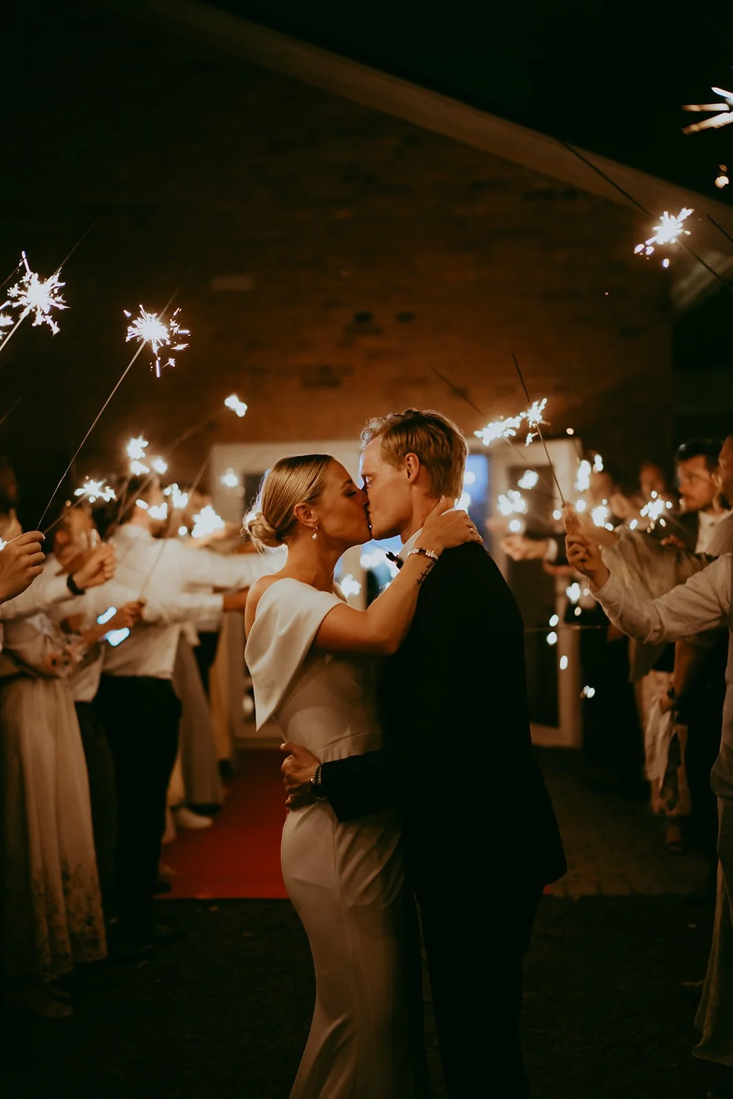 A couple is kissing at a celebration surrounded by people holding sparklers in a warmly lit indoor venue.