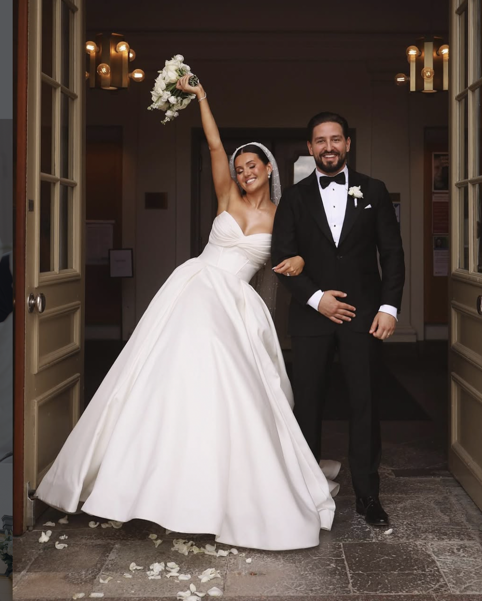 Bride and groom standing at the entrance of a building, celebrating wedding. The bride is holding a bouquet, smiling, with her arm raised. The groom is smiling and dressed in a black tuxedo with a bow tie.