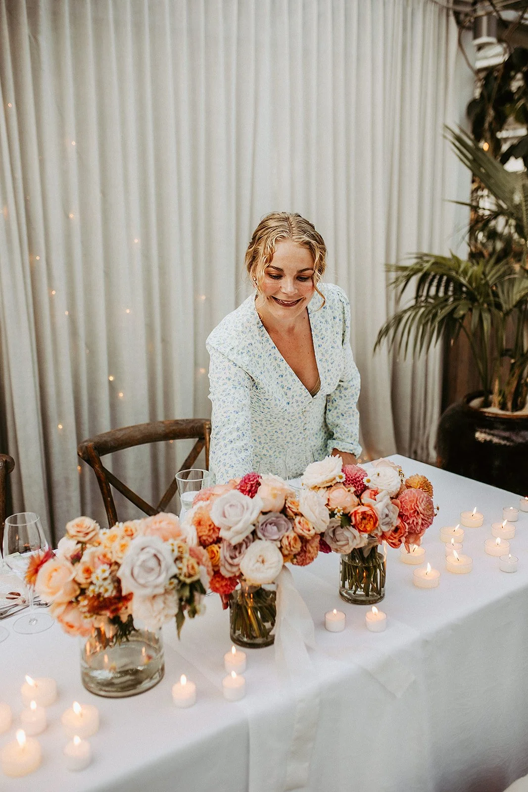 A woman in a light blue floral dress arranging pink and white flowers on a table at a celebration or event, with candles and glassware, and a backdrop of cream curtains and greenery.