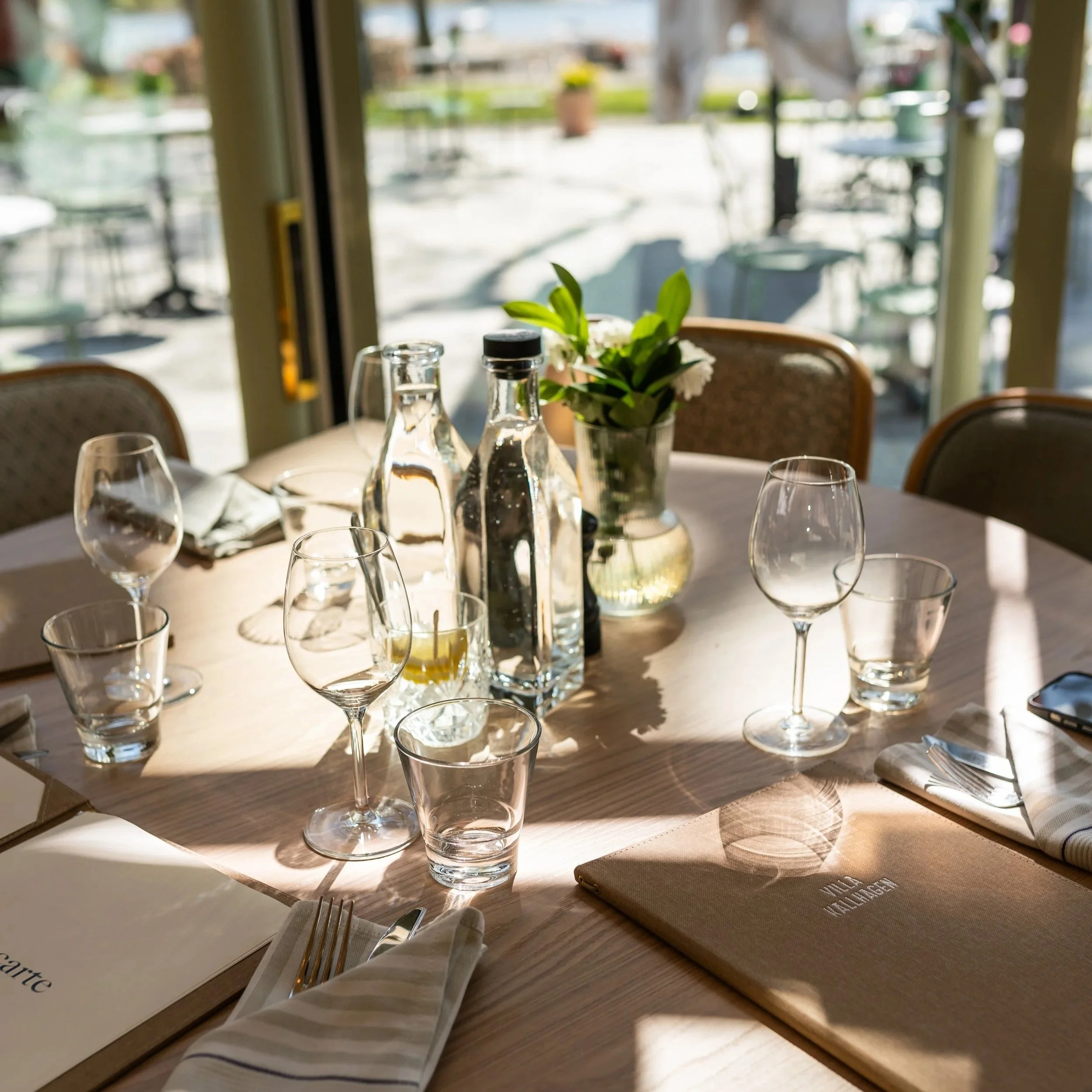 A dining table set with empty wine glasses, a small vase with green leaves and white flowers, and water bottles, near a window with sunlight and blurred outdoor scenery.