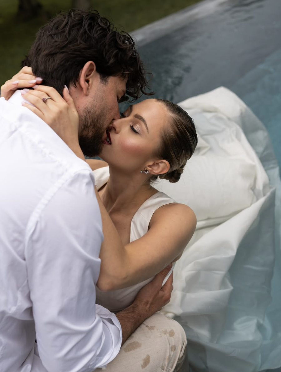 A couple sharing an intimate moment, about to kiss, lying in a bed with white sheets.