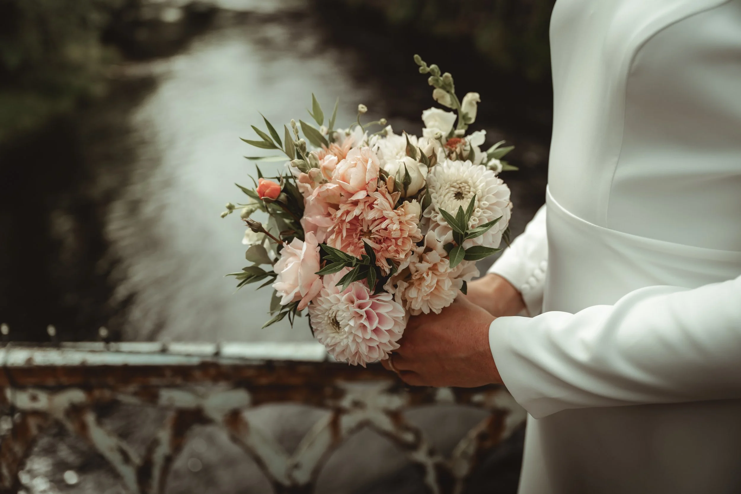 A person in white attire holding a bouquet of pink and white flowers near a river or stream with rocks and water in the background.