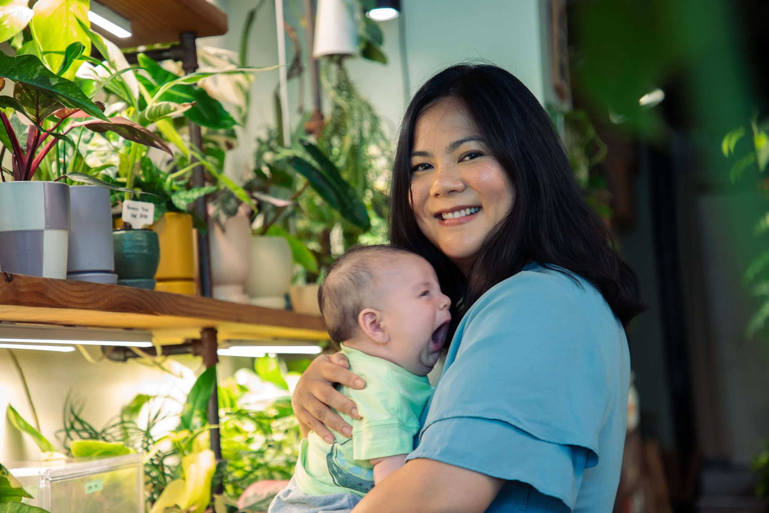 A woman holding a yawning baby in a room with potted plants and wooden shelves.