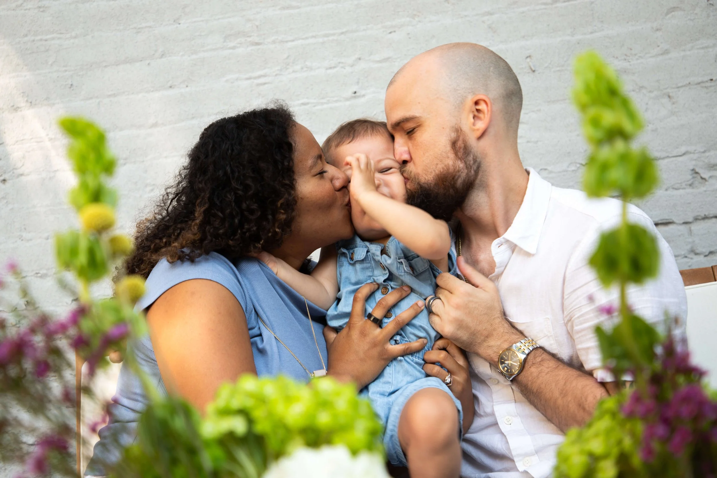 A family of three, a woman, man, and a young child, sharing a tender moment with kisses on the cheeks, surrounded by flowers with a white brick wall background.