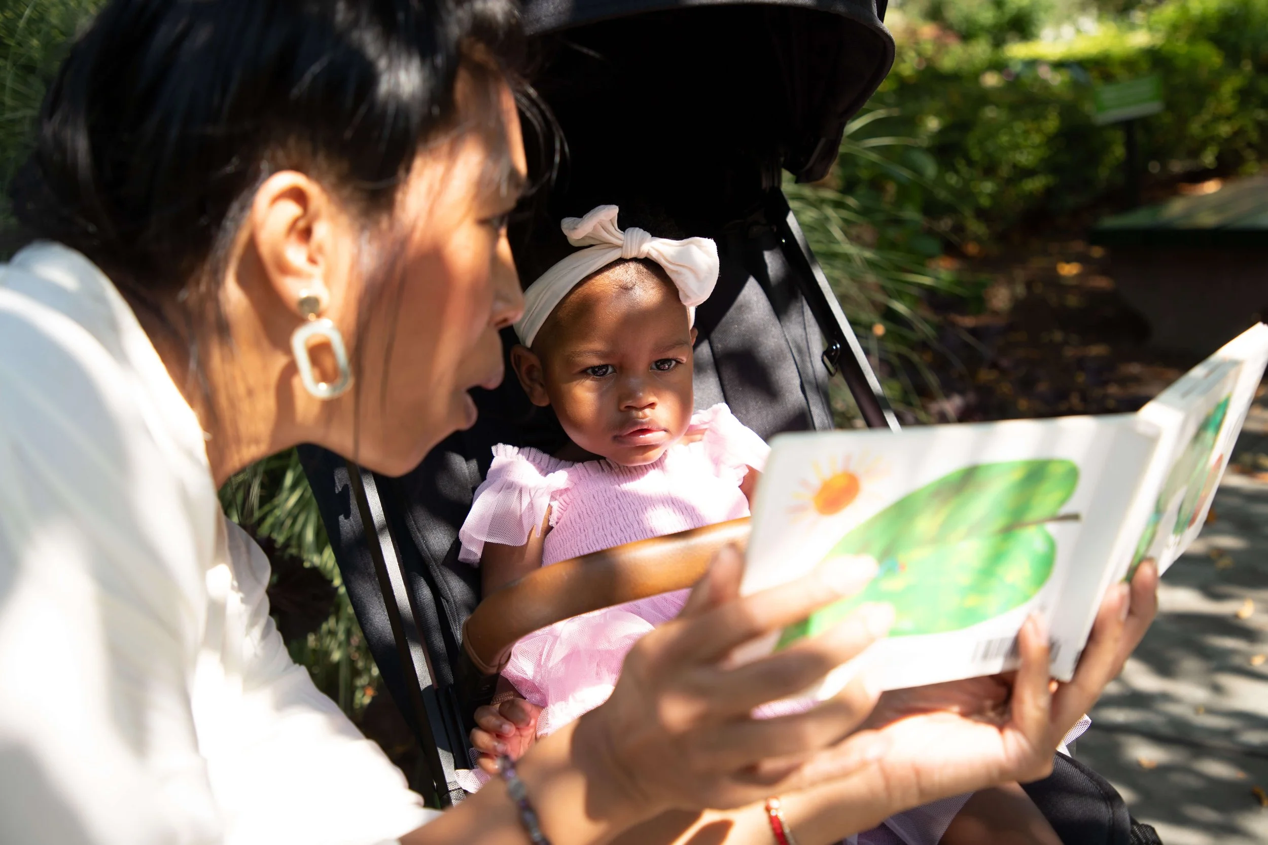 An older woman reading a picture book to a young girl sitting in a stroller outdoors.