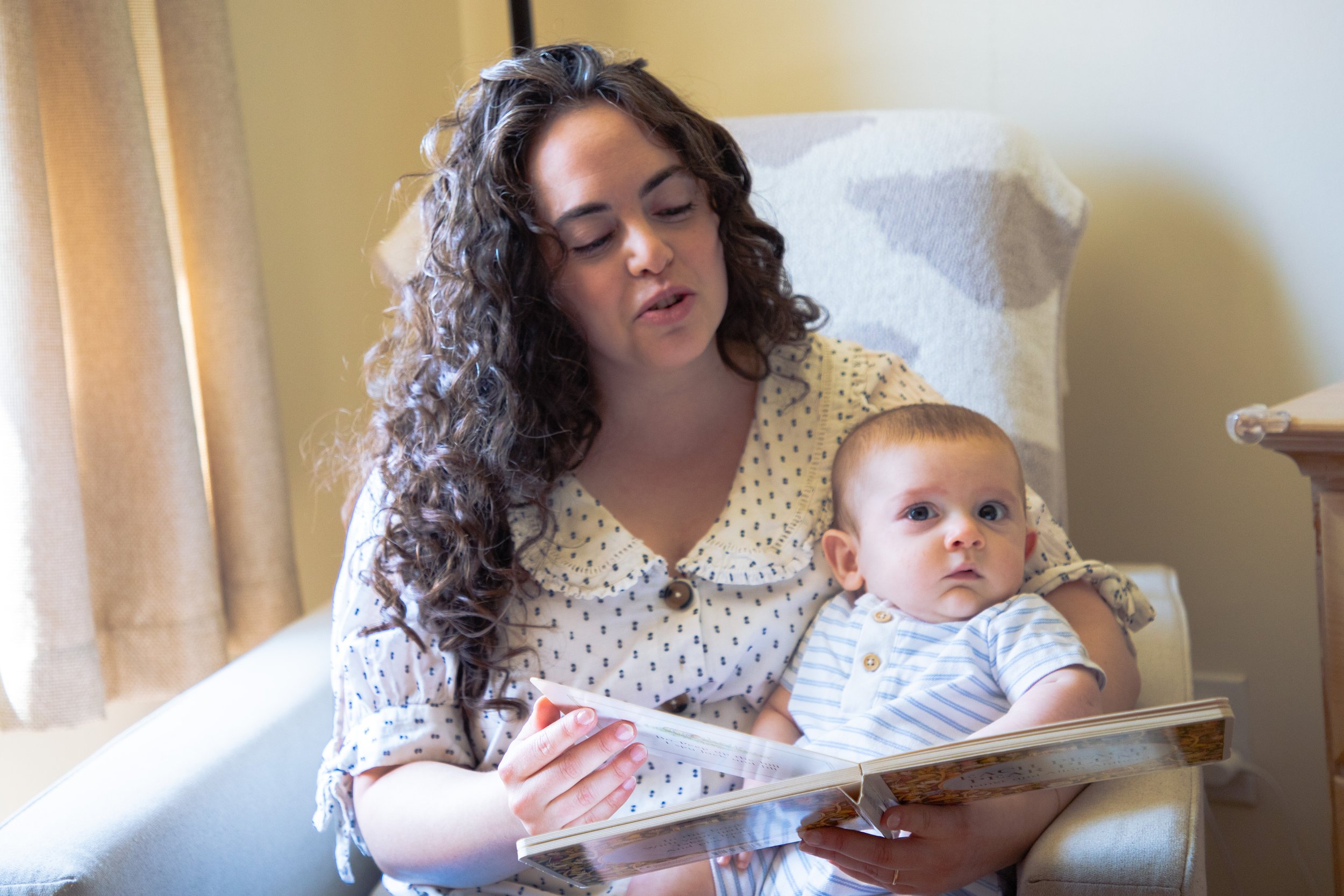A woman with curly brown hair reading a children's book while holding a baby in her lap, seated on a beige armchair in a room with light-colored walls and a window with curtains.