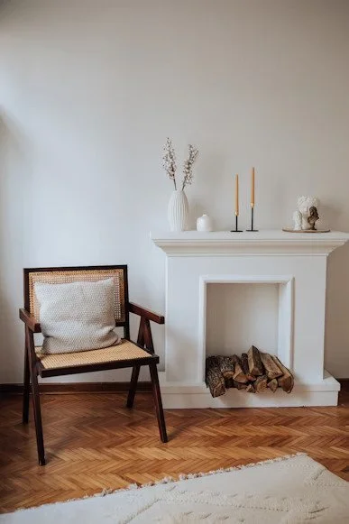 Living room with a wooden armchair, decorative pillow, white fireplace mantel, vase with dried flowers, candles, small sculpture, and stacked logs inside the fireplace.