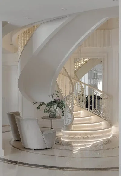 Elegant spiral staircase with a curved white banister and built-in lighting, situated near a seating area with a chair and small table with a potted plant inside a bright, modern home.
