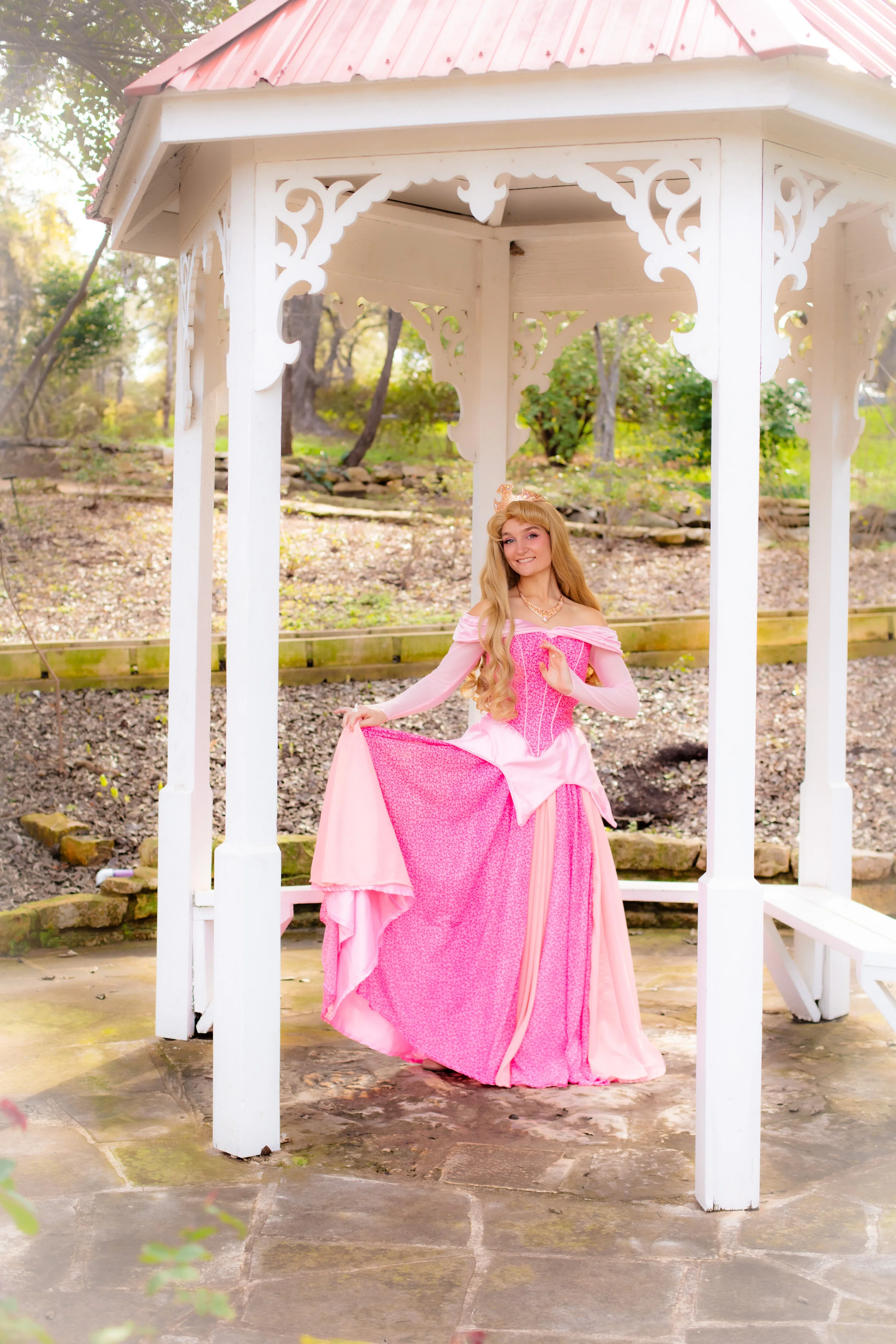 A woman dressed as a princess in a pink gown stands under a white gazebo in a garden, holding up her gown with one hand and smiling.