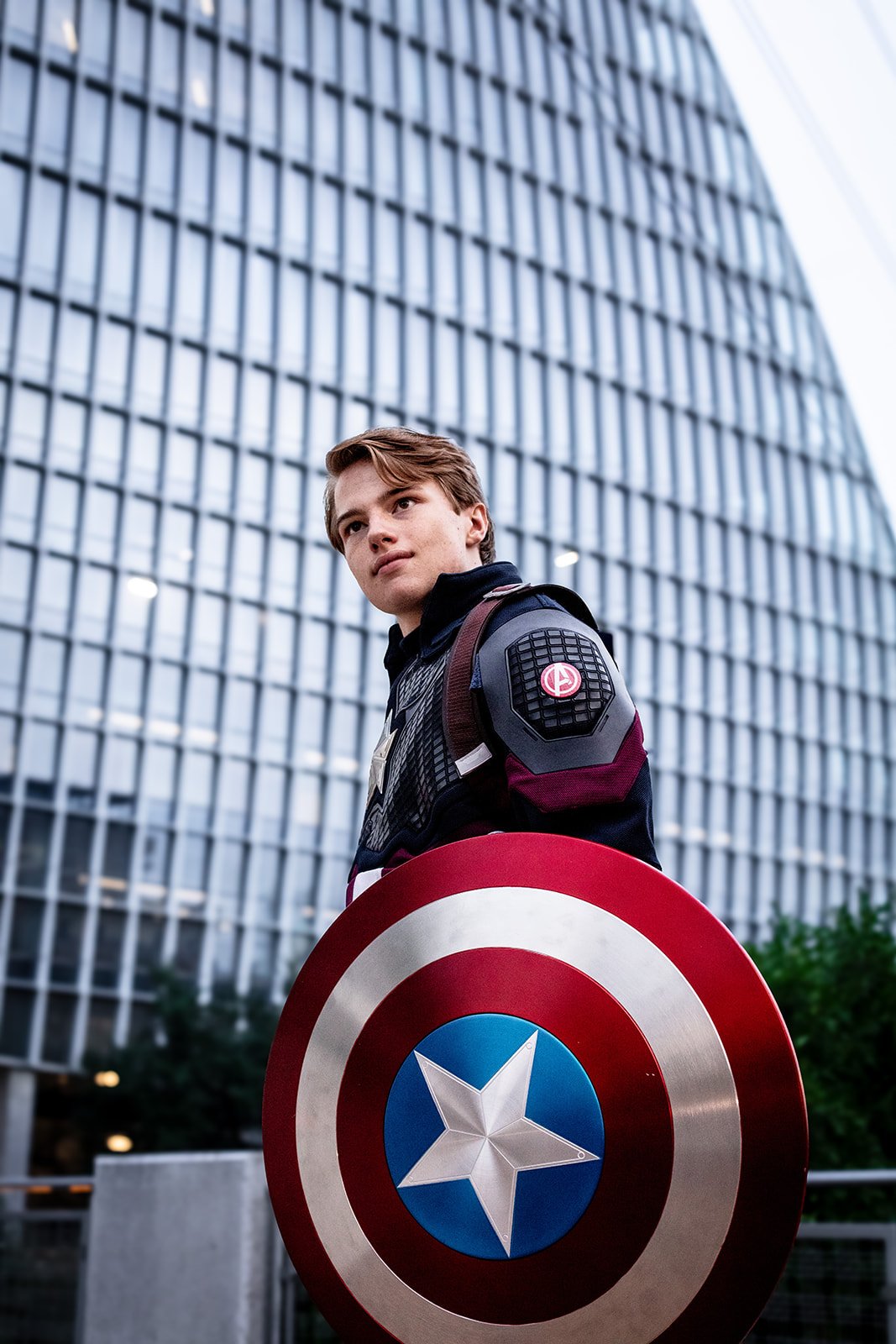 A young man in a Captain America costume holds Captain America's shield in front of a modern glass office building.