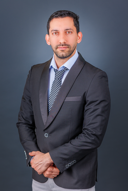 A man with dark hair and a beard wearing a dark gray suit, light blue shirt, and a striped tie, standing against a solid dark background.