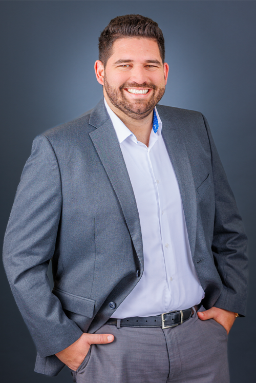 A smiling man in a gray suit and white shirt posing against a dark gray background.