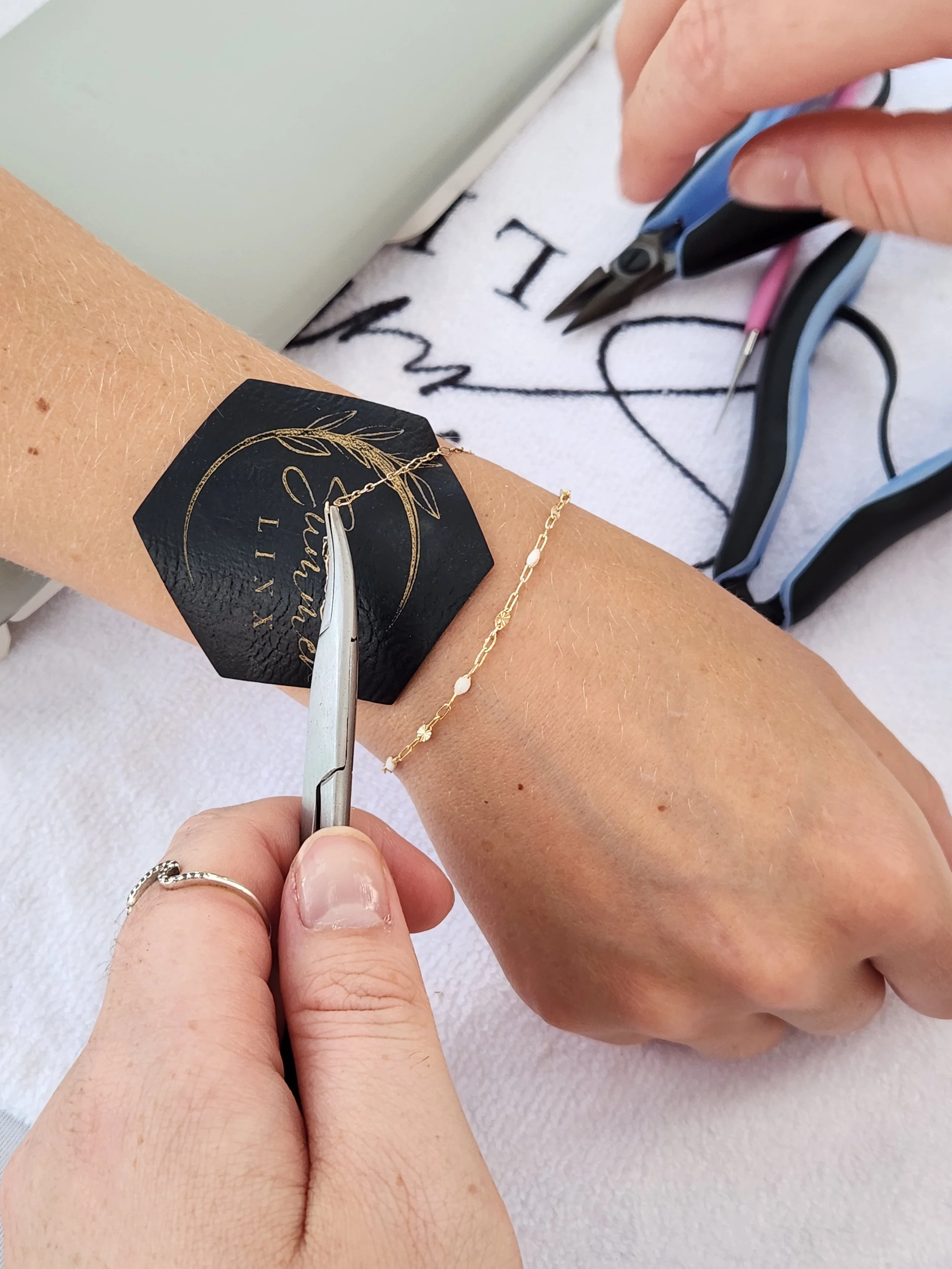 A person receiving a tattoo on their arm, with a tattoo artist holding a tattoo needle and working on the design. The person has a gold bracelet on their wrist and a black wristband with a logo.
