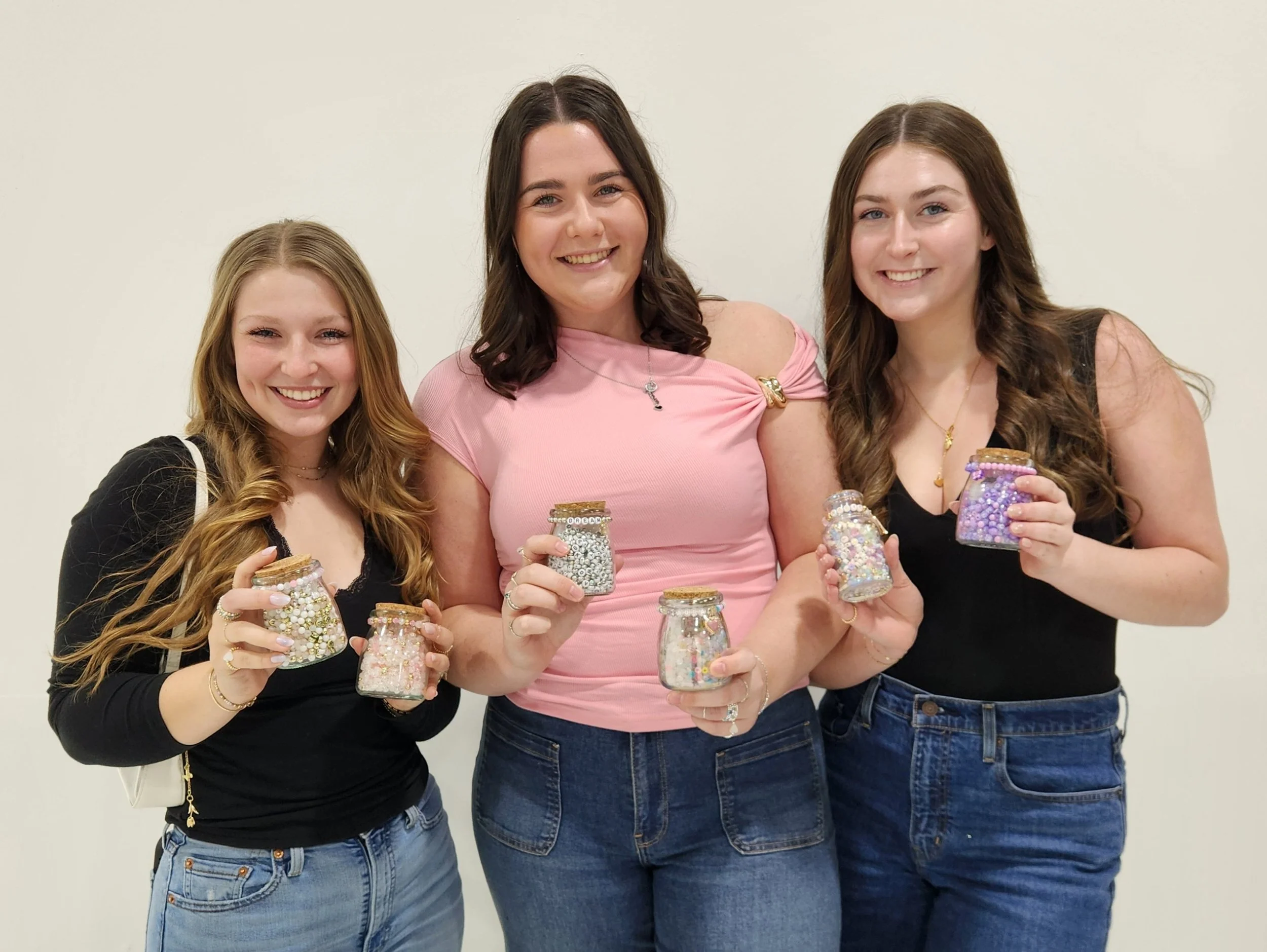 Three young women smiling and holding jars filled with colorful beads or sequins, standing against a plain white wall.