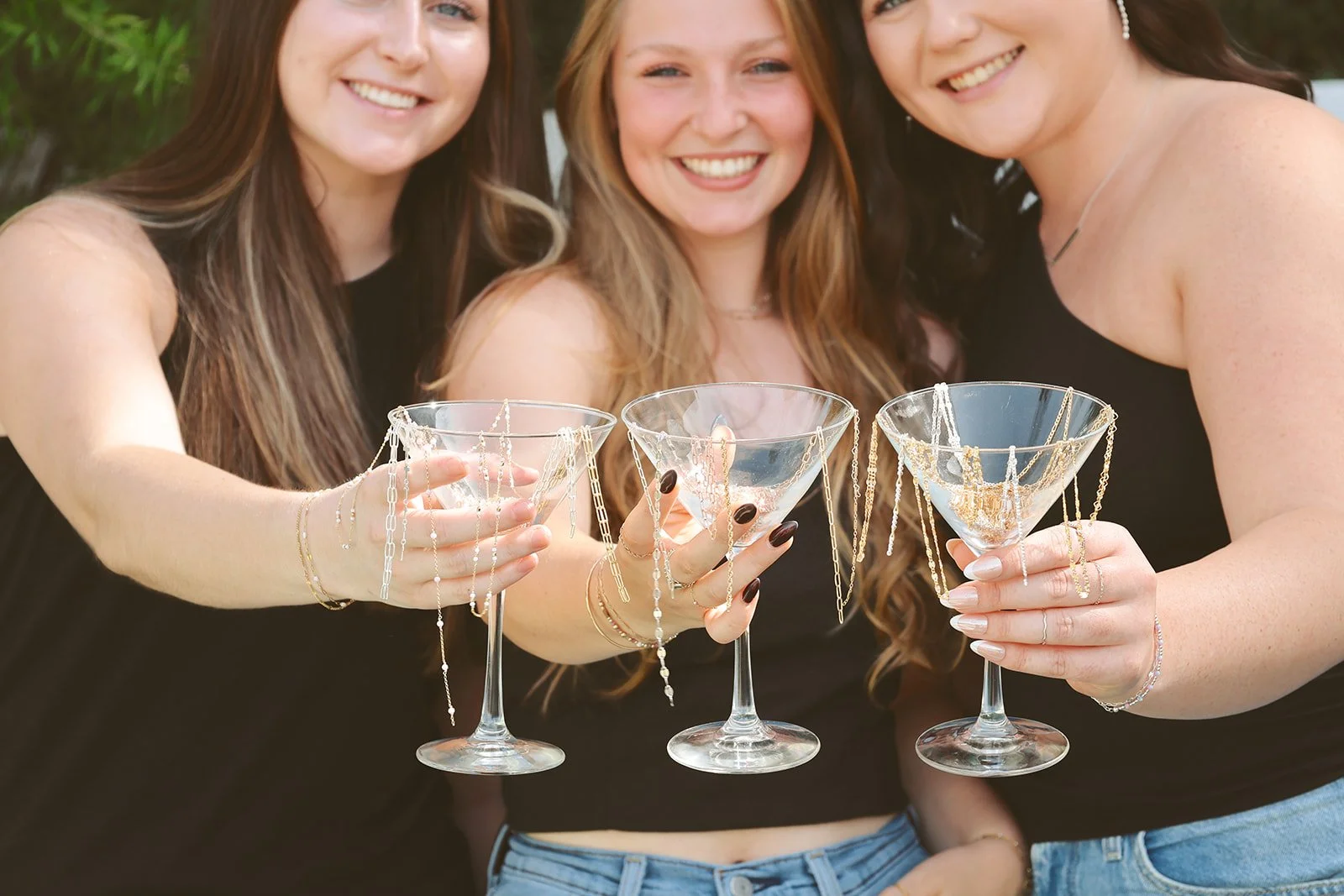 Three women smiling and holding cocktail glasses decorated with jewelry for a celebration.