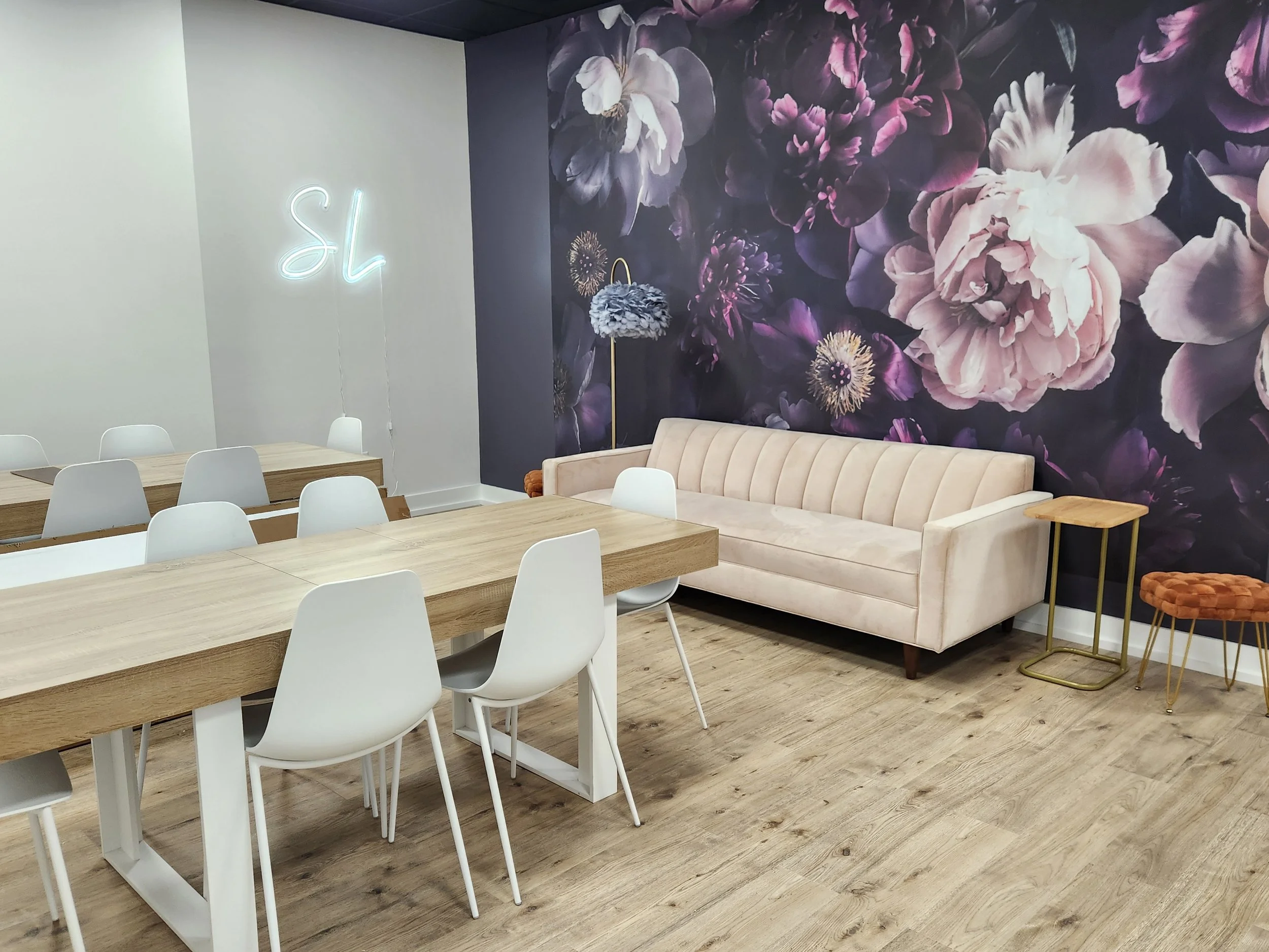 A room with a light-colored velvet sofa, a floral wallpaper wall, a round side table, a decorative lamp with a feather shade, a pig-shaped stool, a long wooden table, and white chairs. There's also a neon sign on the wall that reads 'sl.'